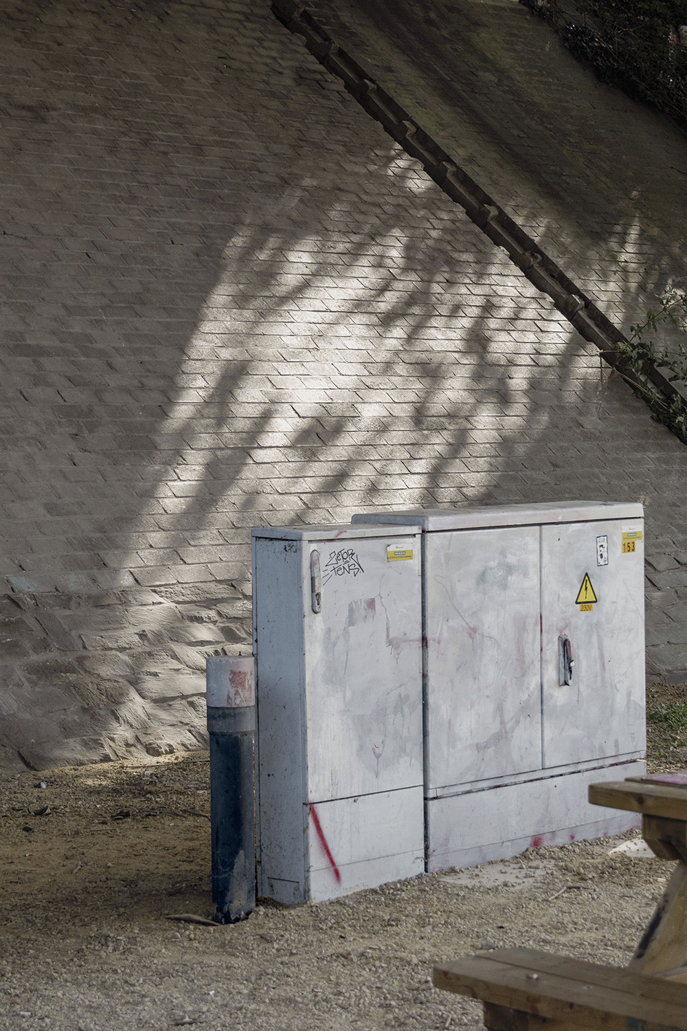 Electrical box on the ground near a brick wall with shadows of tree branches and leaves cast on it. A bench is partially visible in the foreground.