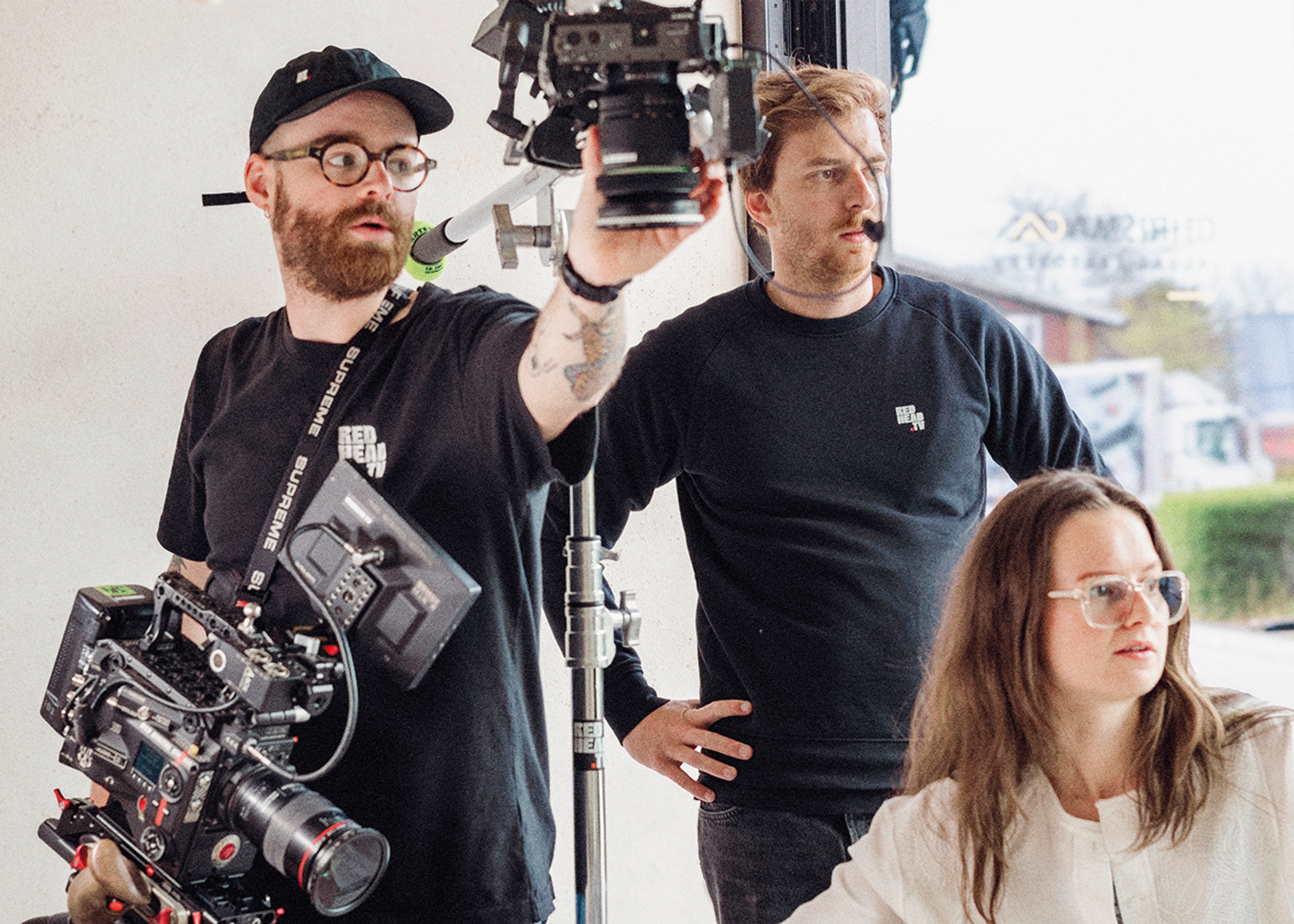 Two men on a film set working with camera equipment, one in black shirt and glasses with a beard, and the other with reddish hair and a black shirt, along with a woman with brown hair and glasses, during daytime.