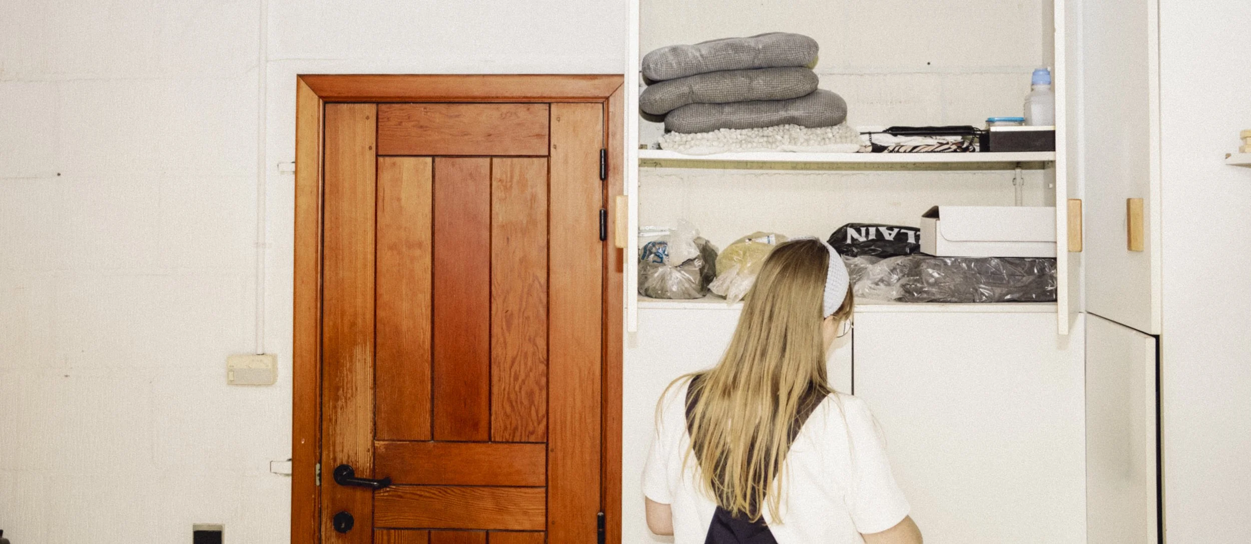 A woman with long blonde hair wearing a white shirt and headband is seen from behind, facing a white storage cabinet filled with folded gray towels, plastic bags, and boxes, next to a wooden door with a black handle.