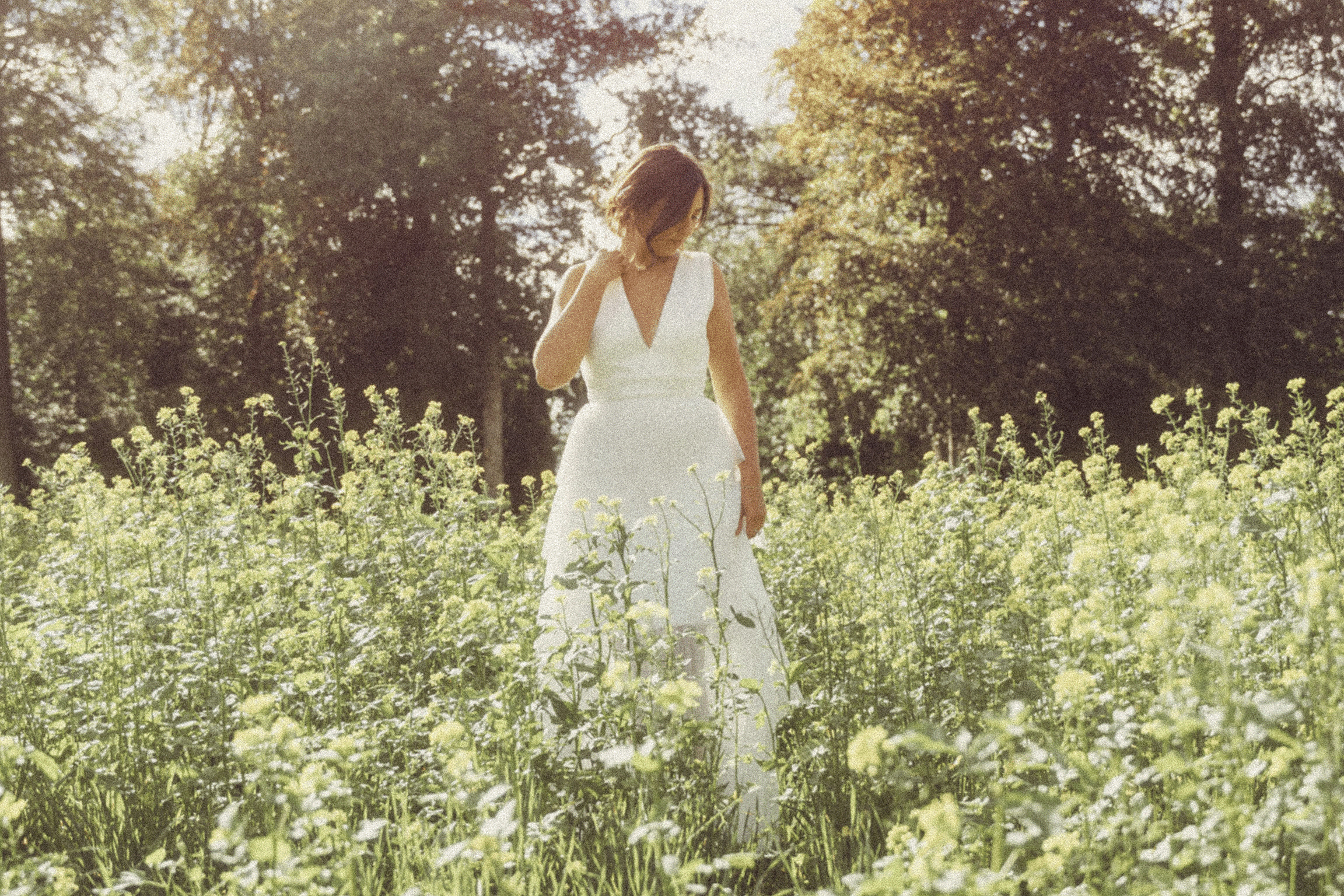 A woman in a white dress walking through a field of yellow flowers with trees in the background, illuminated by sunlight.
