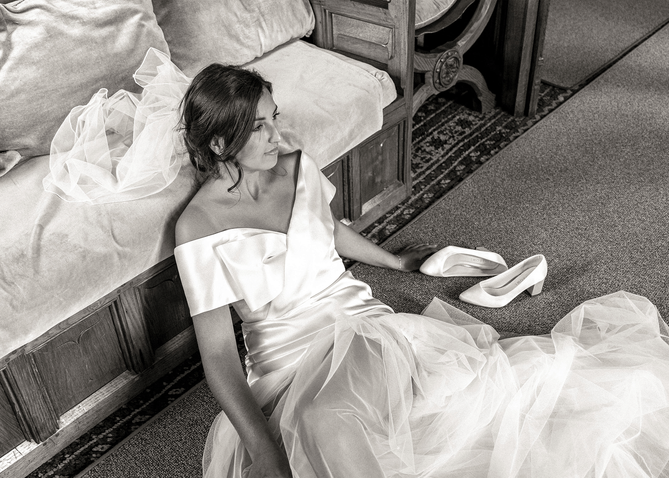 A woman in a wedding dress lying on a bed, with white high-heeled shoes placed on the floor nearby.