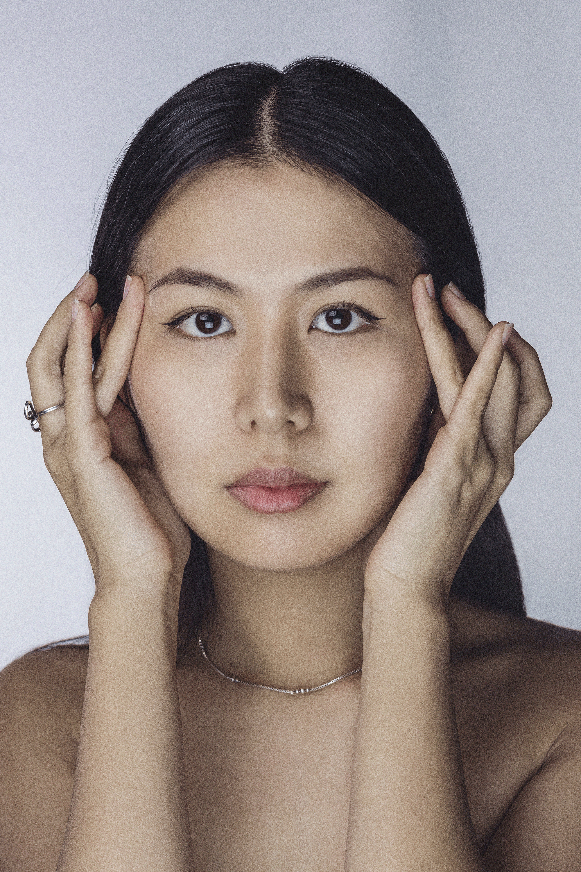 Close-up of a woman's face with her hands near her face, dark hair, light makeup, and a neutral background.