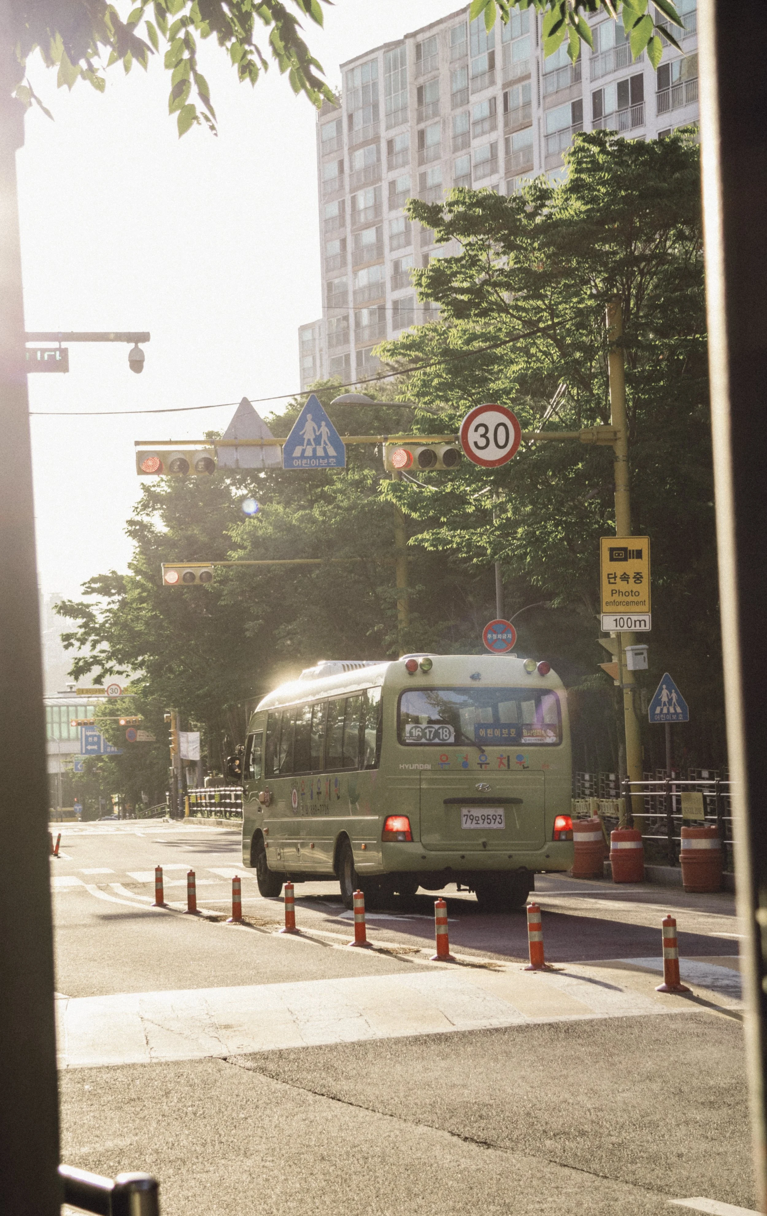City street scene with a small green bus, traffic cones, traffic lights, and road signs, including a speed limit of 30 km/h and a pedestrian crossing sign. Tall buildings and trees are in the background, and sunlight shines through the scene.