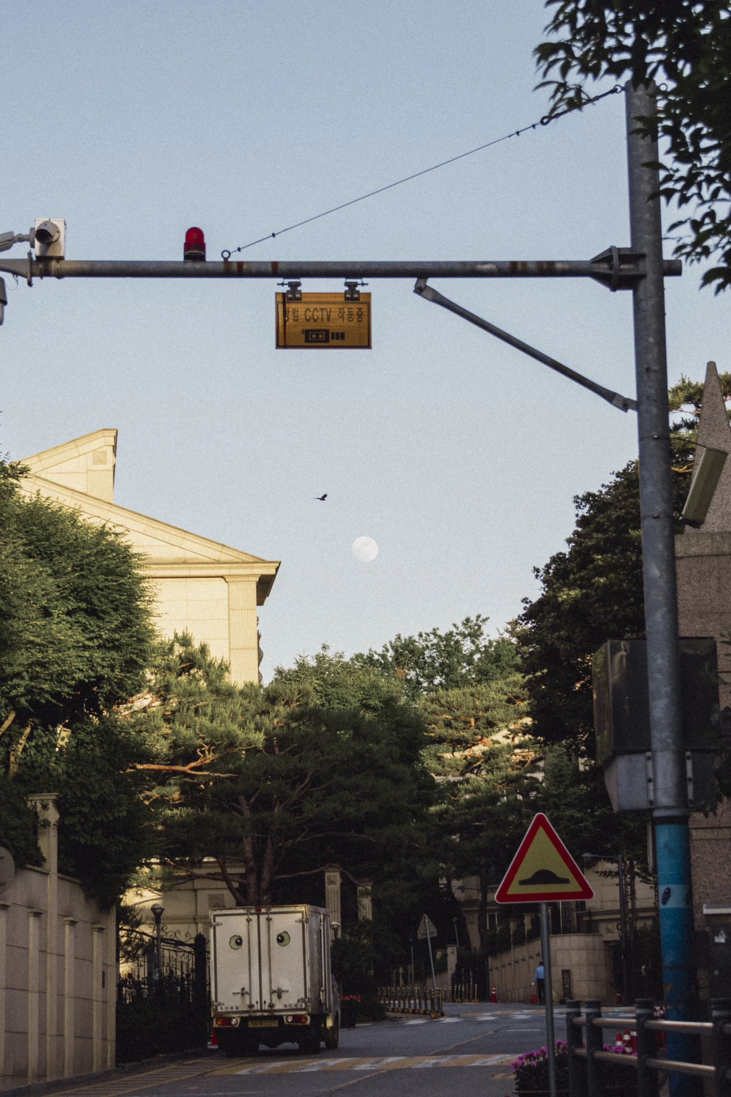 City street scene at dusk with trees, a building, traffic signs, and a delivery truck on the road. The moon is visible in the sky.