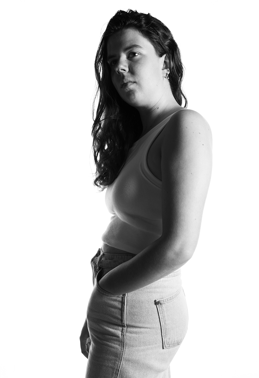 Black and white portrait of a woman with long wavy hair, wearing a tank top and jeans, looking to her left with her hand in her pocket.