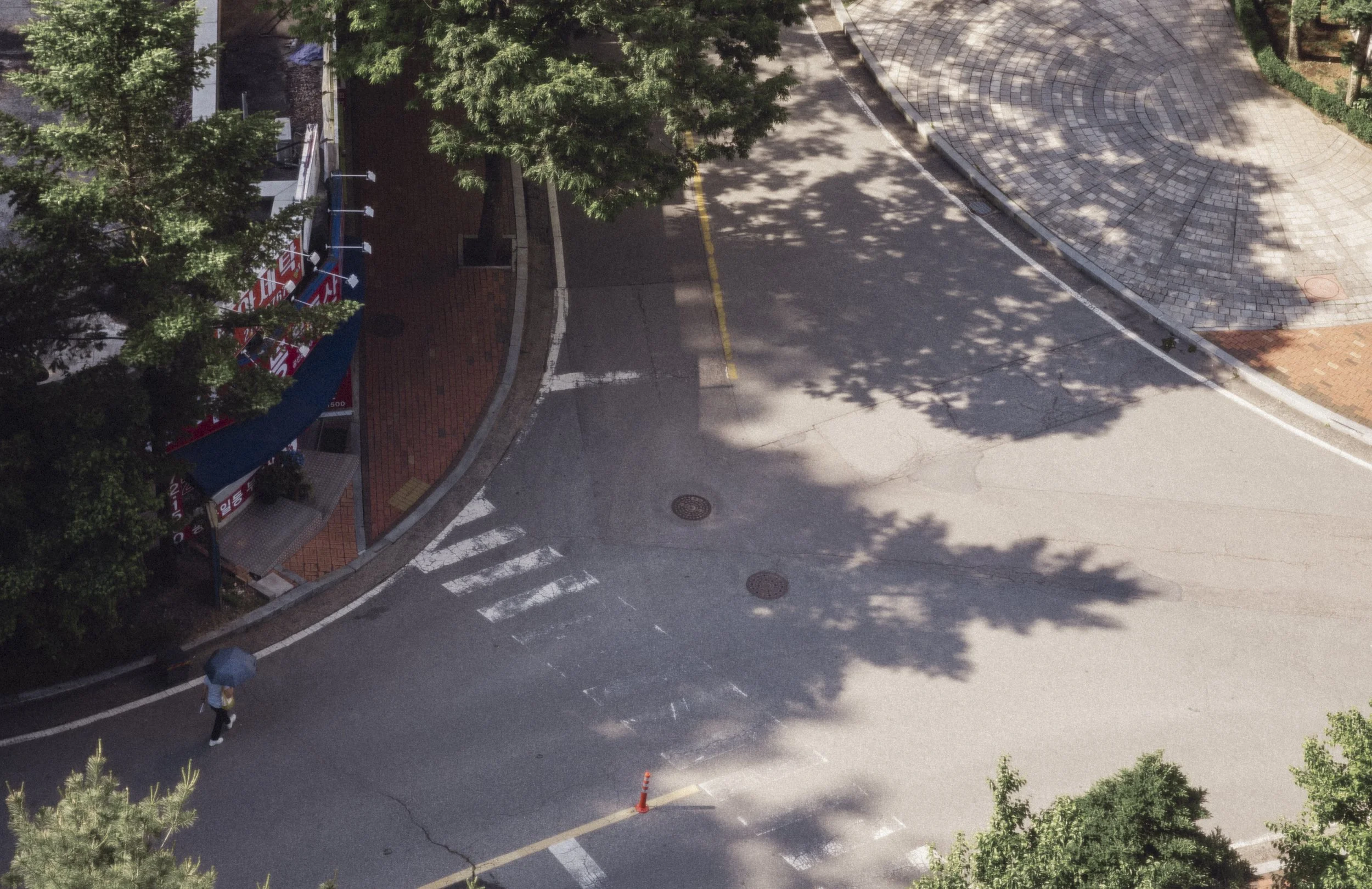 An aerial view of an empty street with a sidewalk and trees casting shadows, a person walking with a blue umbrella, and a traffic cone on the road.