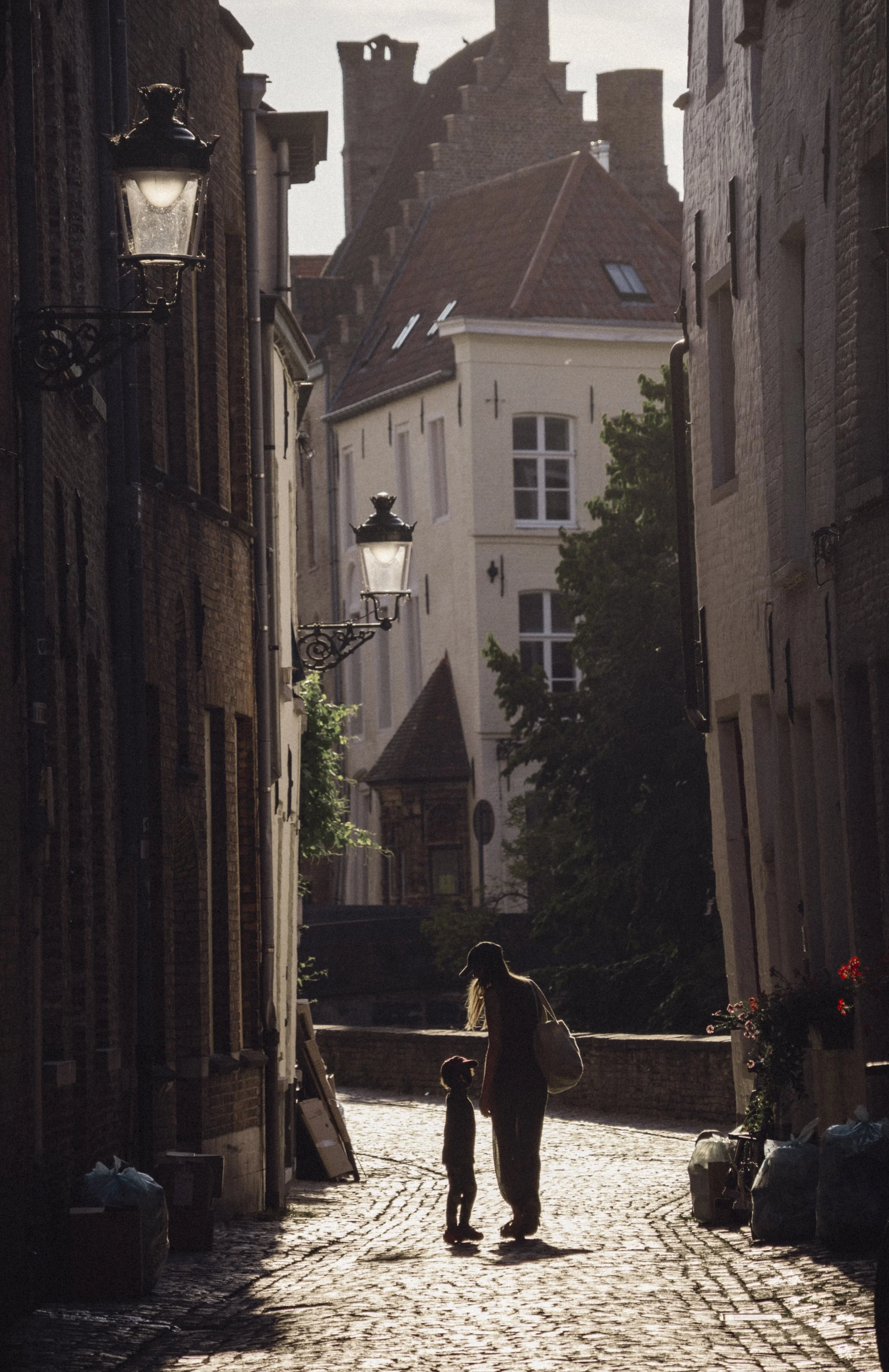 Silhouetted woman and child walking on cobblestone street in alleyway at sunset, with historic buildings, street lamps, and tree in the background.