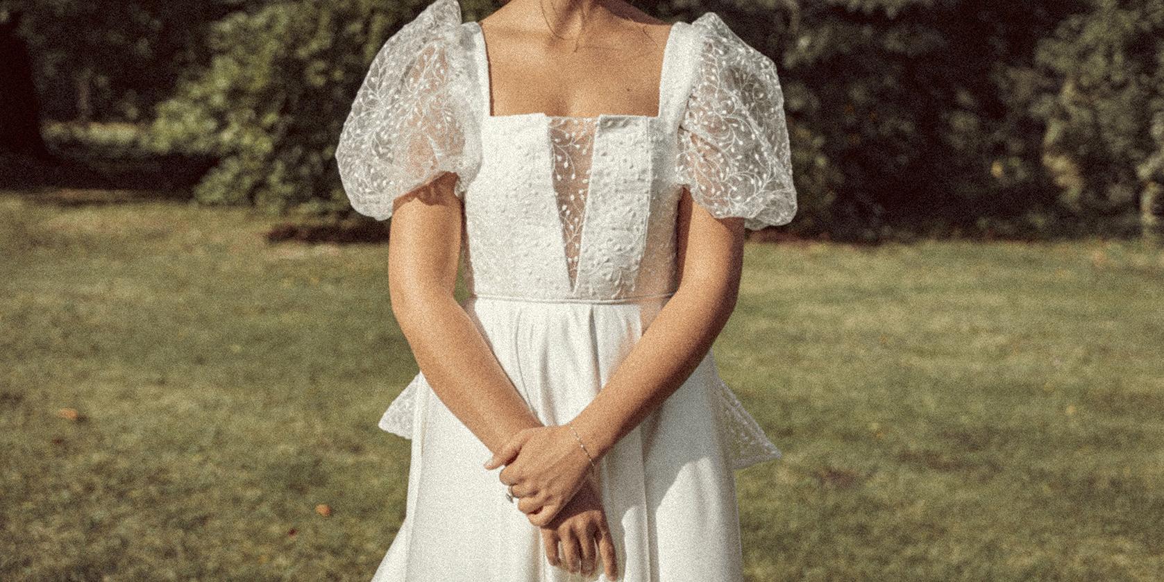 A woman standing outdoors in a garden, wearing a white dress with puffed lace sleeves and a deep V-neckline, with her hands clasped in front of her.
