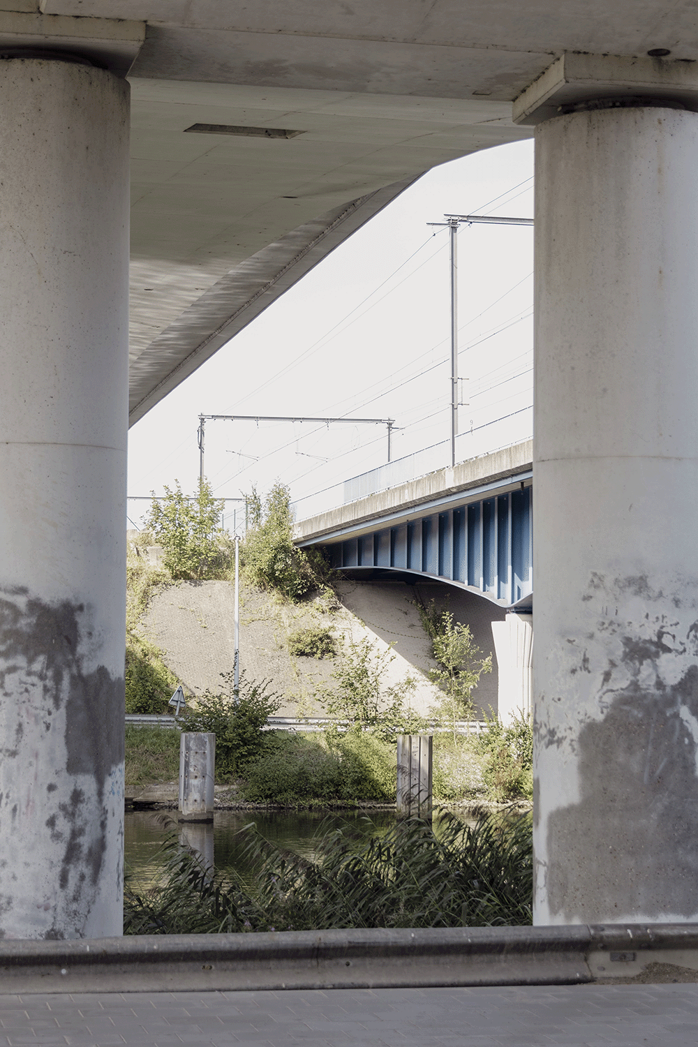 View of an overpass bridge from beneath, showing large supports, the underside of the bridge, and electrical lines above. There are trees and bushes along the embankment with a small waterway underneath.