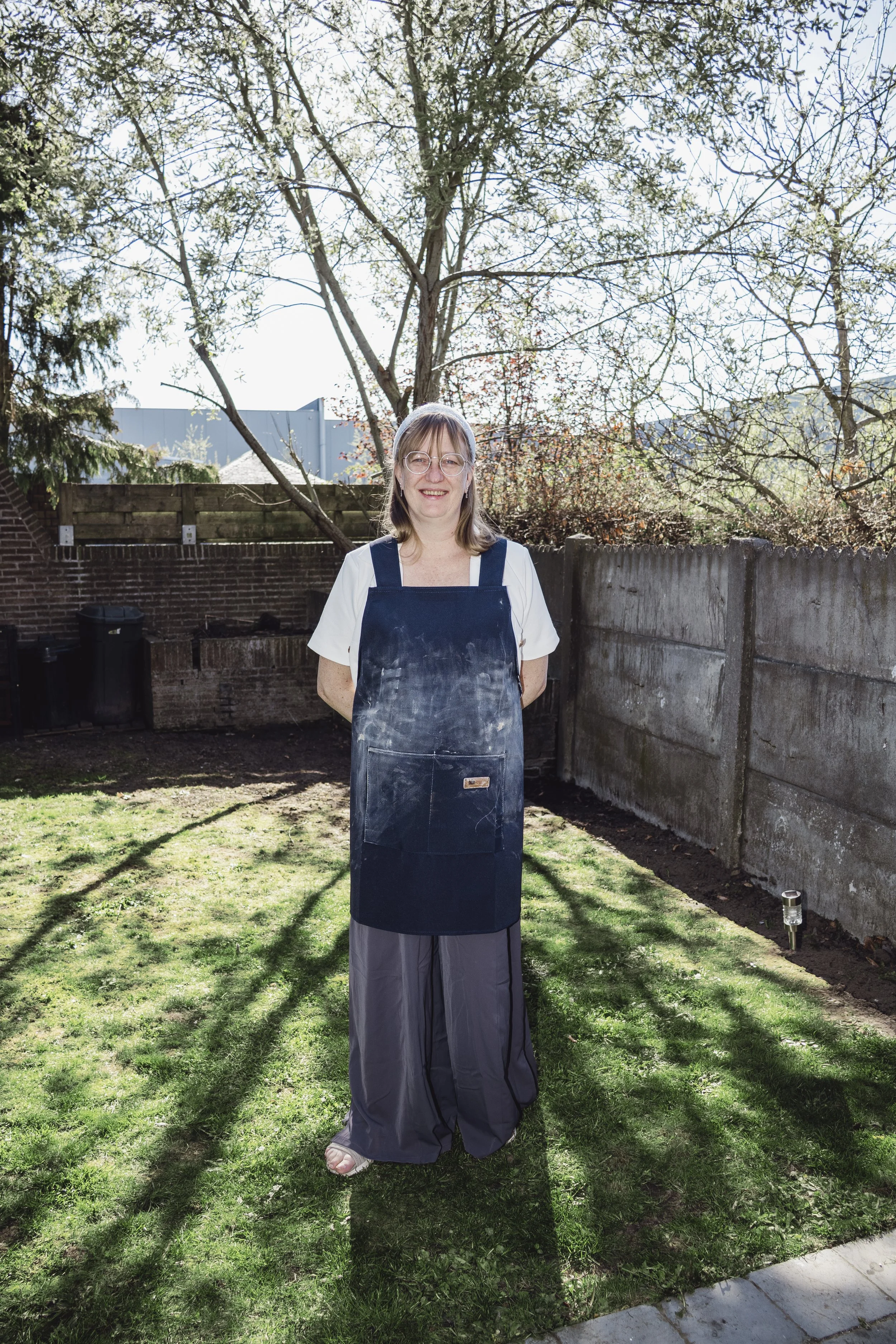 A woman standing outdoors on a grassy area with trees and a brick wall behind her, wearing glasses, a white shirt, and a black apron, smiling at the camera.