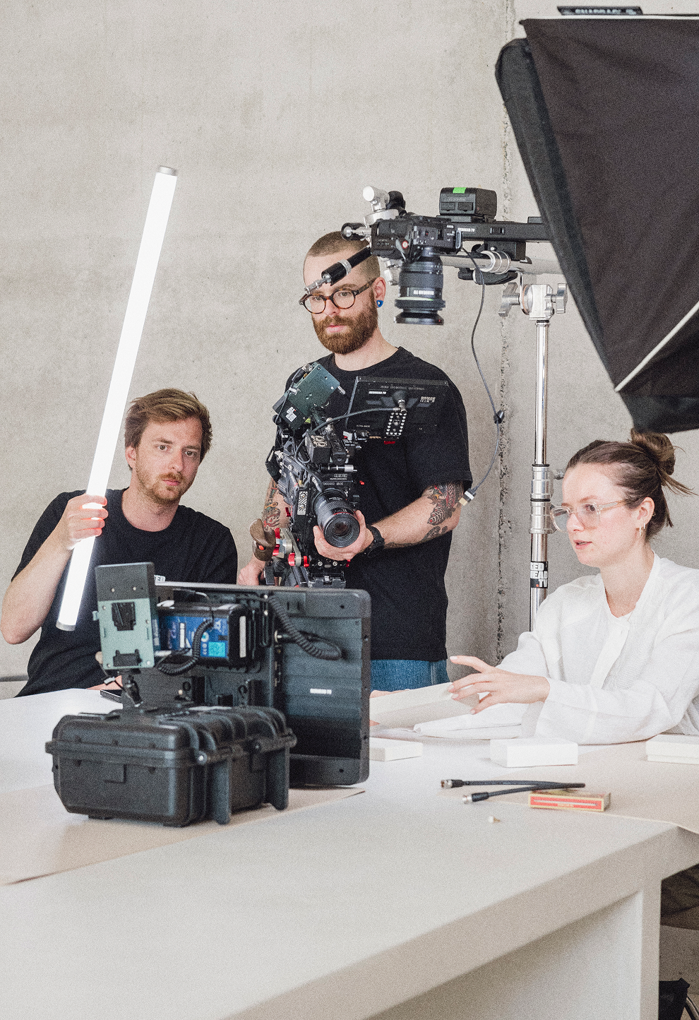 People working on a film set with a large camera, lighting equipment, and a woman sitting at a table with papers.
