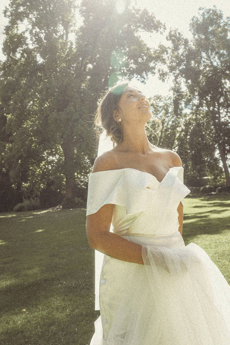 A woman in an off-the-shoulder white dress standing outdoors on a sunny day, smiling and looking up at the sunlight with trees in the background.