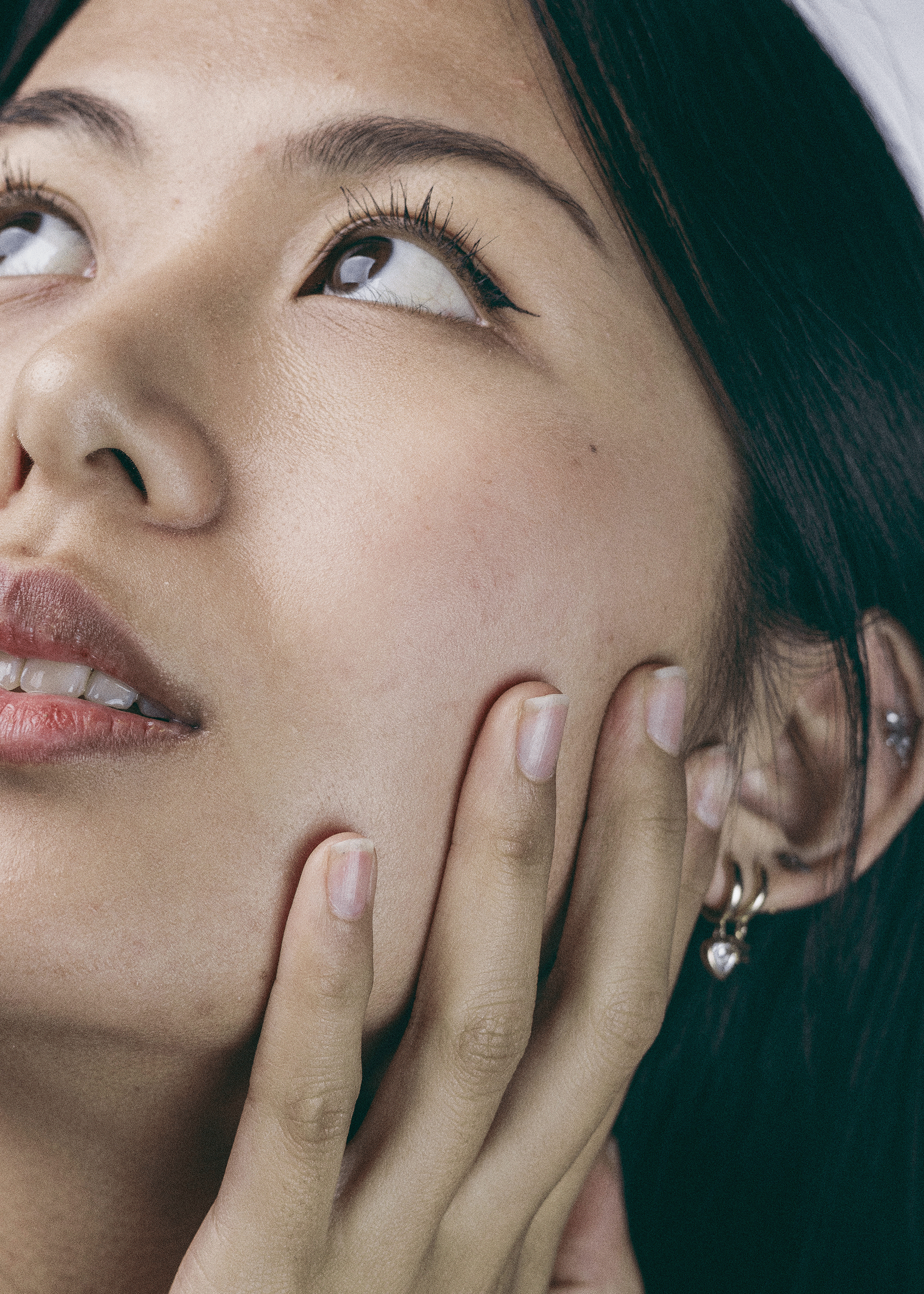 Close-up of a person with makeup, wearing earrings, posing with fingers touching face, hair partially covered by a white headband or cap.