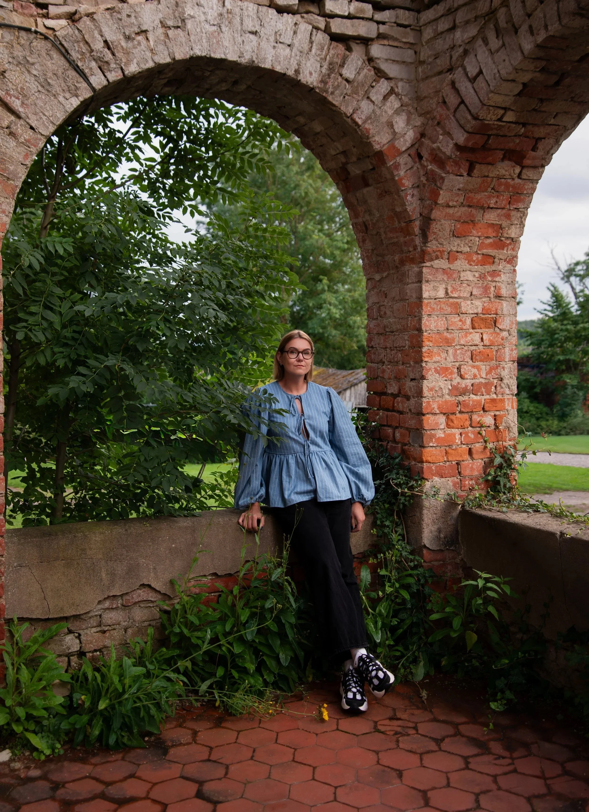 Woman standing outdoors beneath a brick archway, surrounded by green foliage and plants, wearing glasses, a blue striped blouse, black pants, and sneakers.
