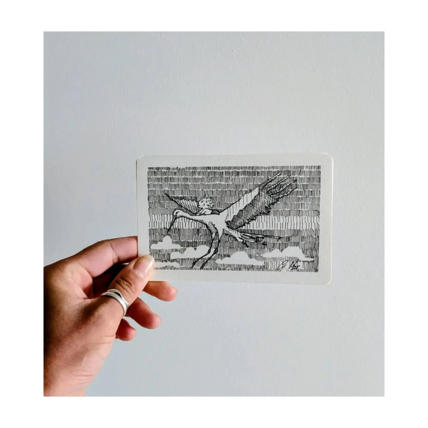 Hand holding a black and white illustration of a human-like figure with wings and a crown, holding a spear, against a brick wall background.