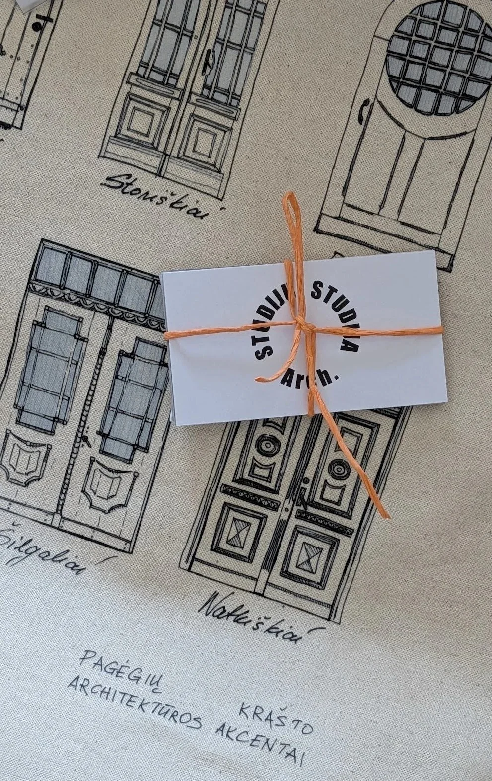 Architectural drawings of store doors and windows with labels, covered by a white card tied with an orange string that reads "STUDIO STUDIO Arch."
