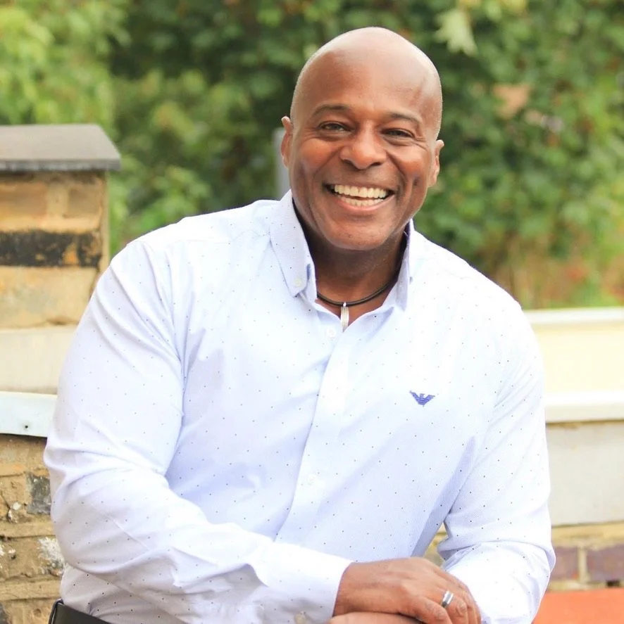 A smiling man with a shaved head wearing a white button-up shirt with small dots, a black choker necklace, and a wedding band, sitting outdoors with greenery in the blurred background.