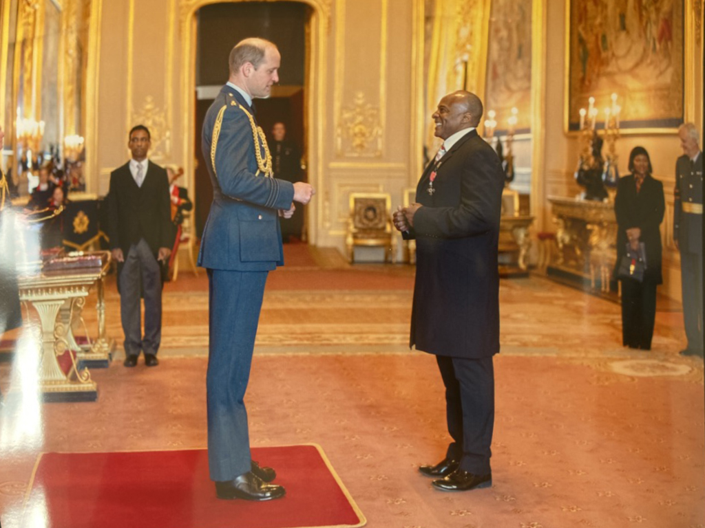 A formal award or medal ceremony taking place in an ornate, gold-accented room with several people in formal attire, including a man in a military uniform and a man in a suit exchanging a medal or award.