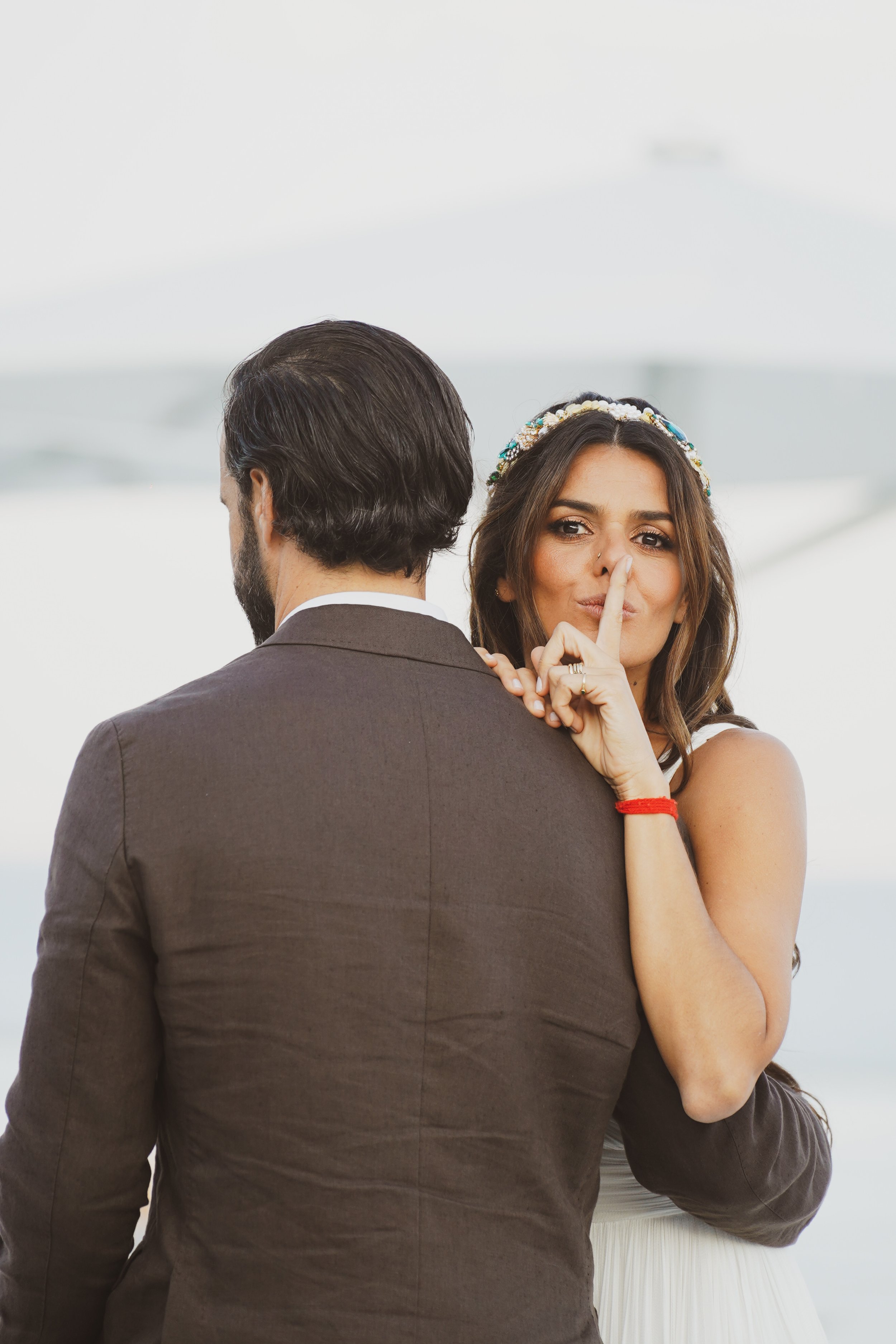 A woman with long brown hair wearing a white dress and colorful headband is holding her finger to her lips, signaling to keep quiet. A man with dark hair and a beard, dressed in a suit, is standing with his back turned, facing away. The background appears to be an indoor or outdoor bright, neutral space.