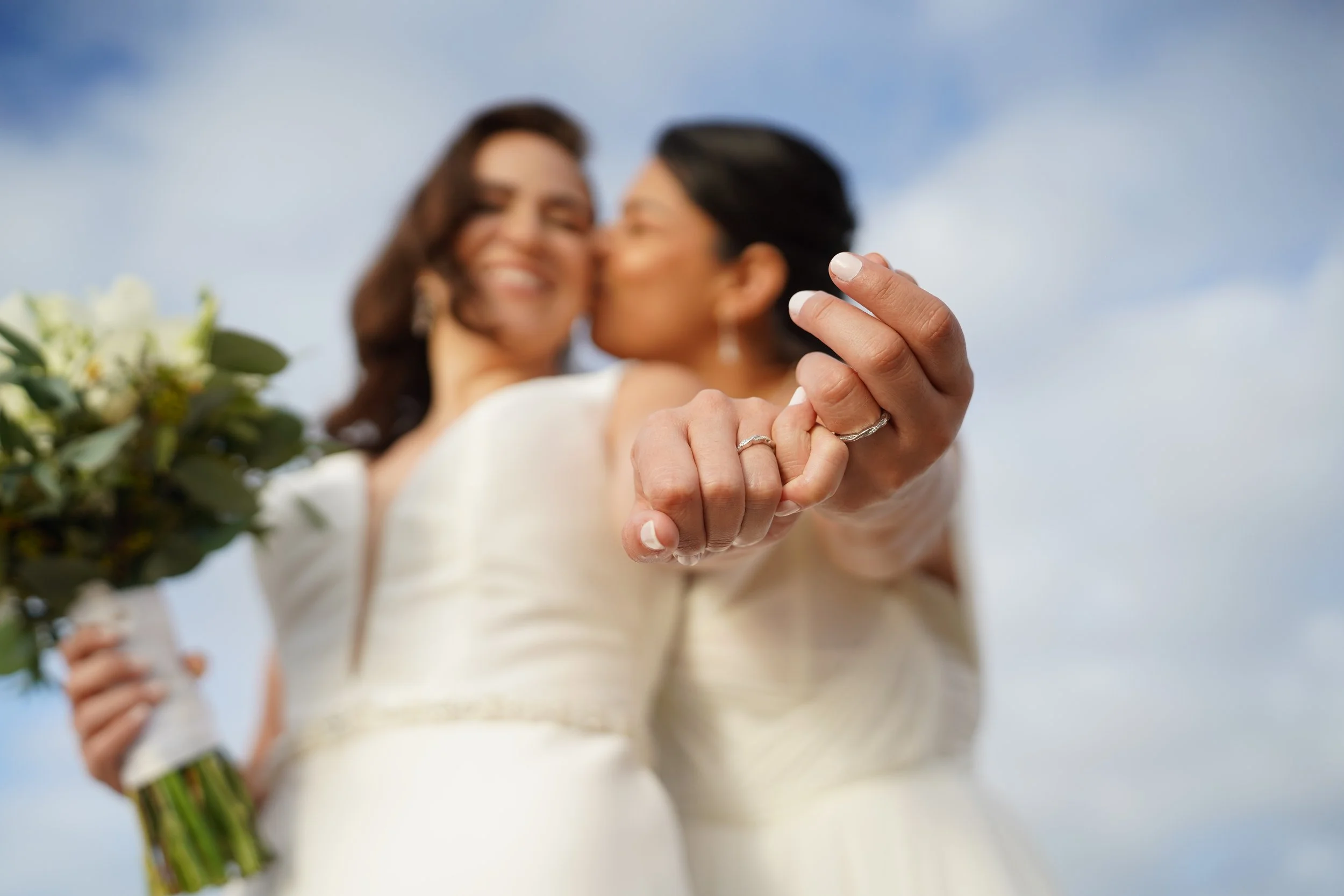 A bride and groom sharing a kiss, with the bride holding a bouquet of flowers, viewed from below with a background of blue sky and clouds.