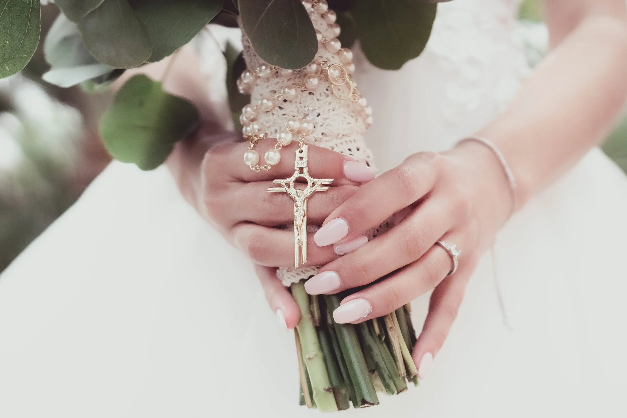 Close-up of a person's hand holding a bouquet of greenery and white flowers, with a gold crucifix and rosary beads hanging from the wrist, person wearing a white dress and a delicate bracelet.