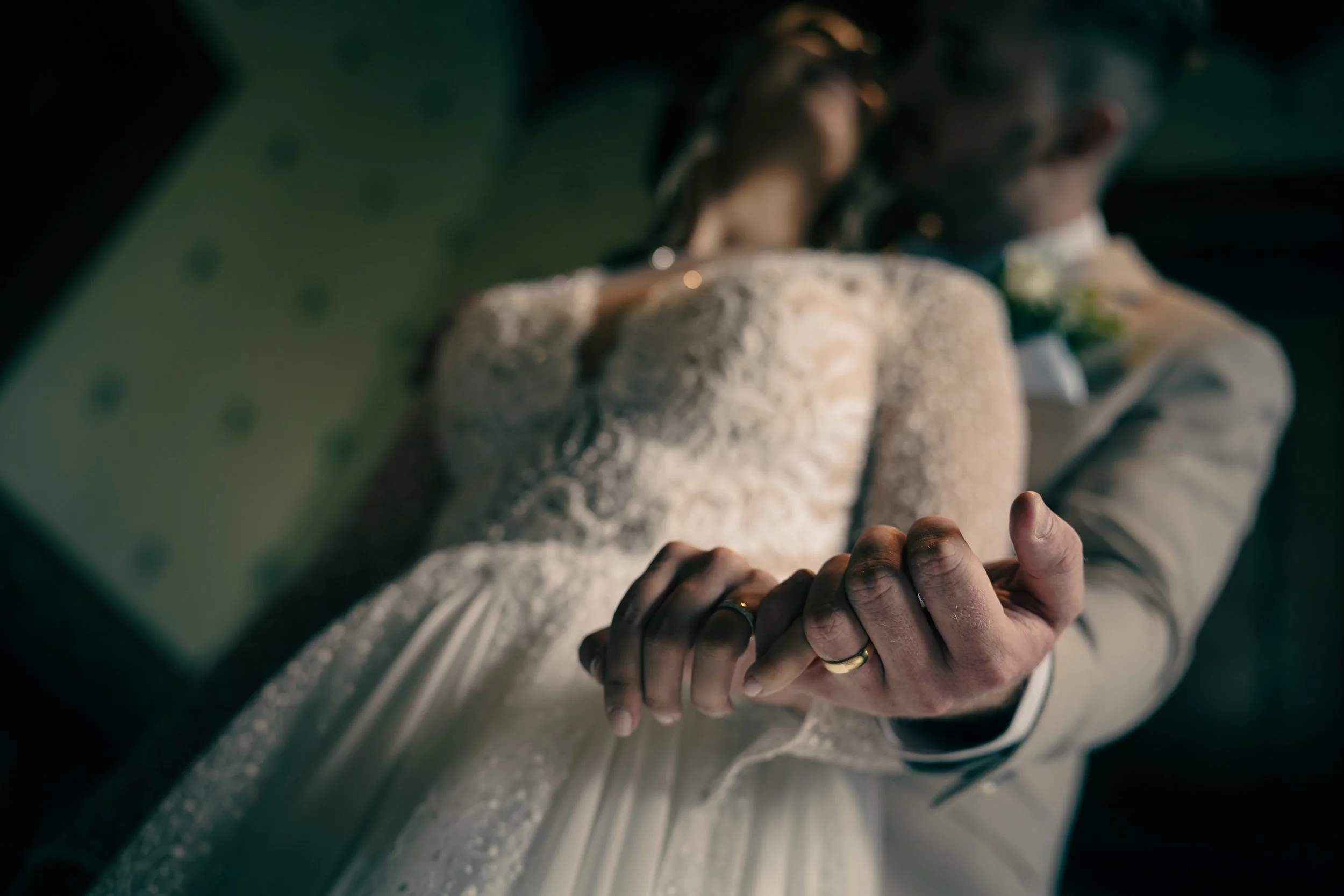 Close-up of a person with wet hands holding onto a metallic surface, possibly a car door handle, with another individual in the background wearing a white characteristic outfit, possibly a wedding dress, and a dark-colored jacket, in a dimly lit setting.