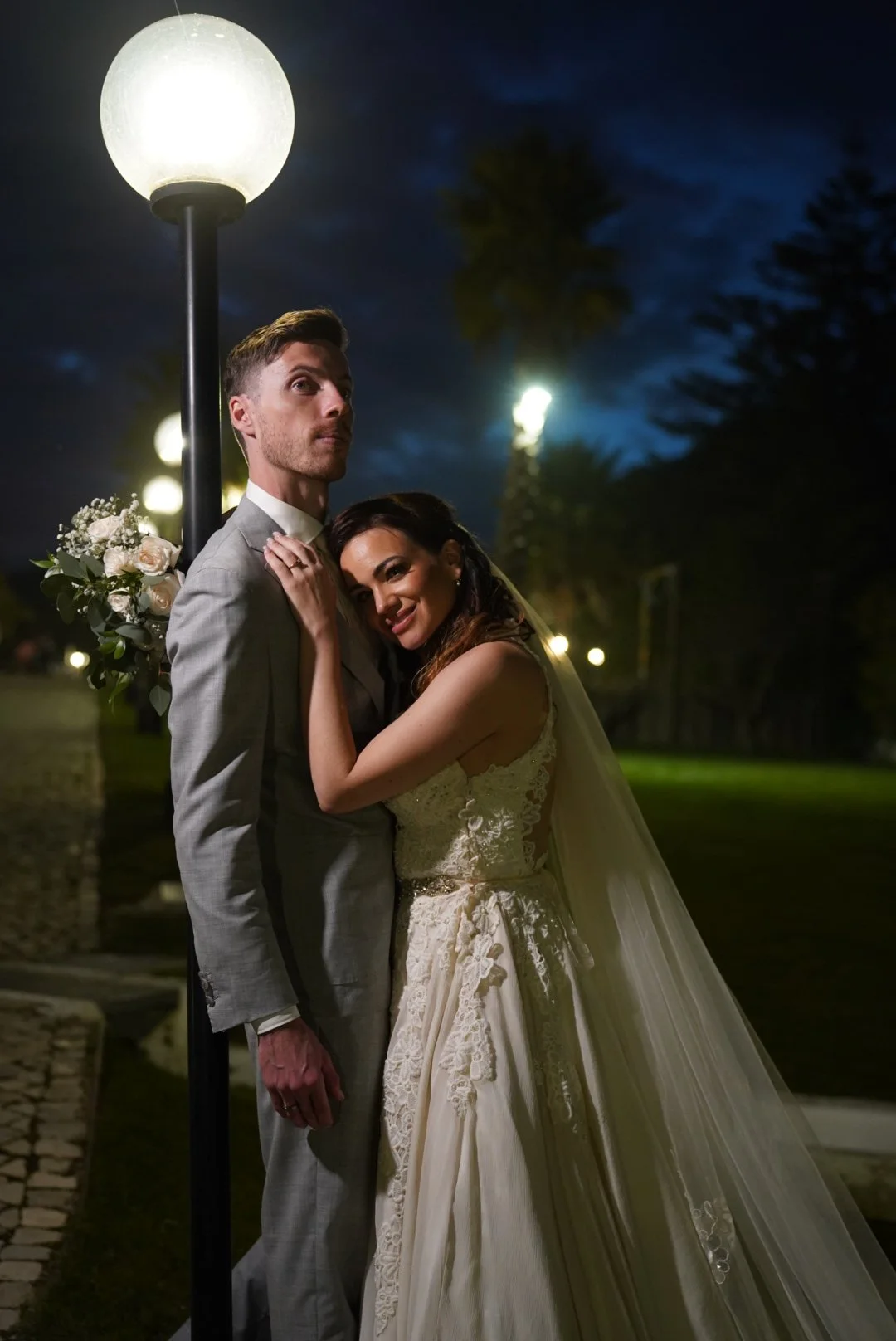 A newlywed couple standing close together at night, illuminated by a streetlamp. The bride is wearing a lace wedding dress with a veil, and the groom is wearing a light gray suit. The bride is smiling and leaning her head on the groom's shoulder, holding his arm, with a bouquet of flowers visible behind him.