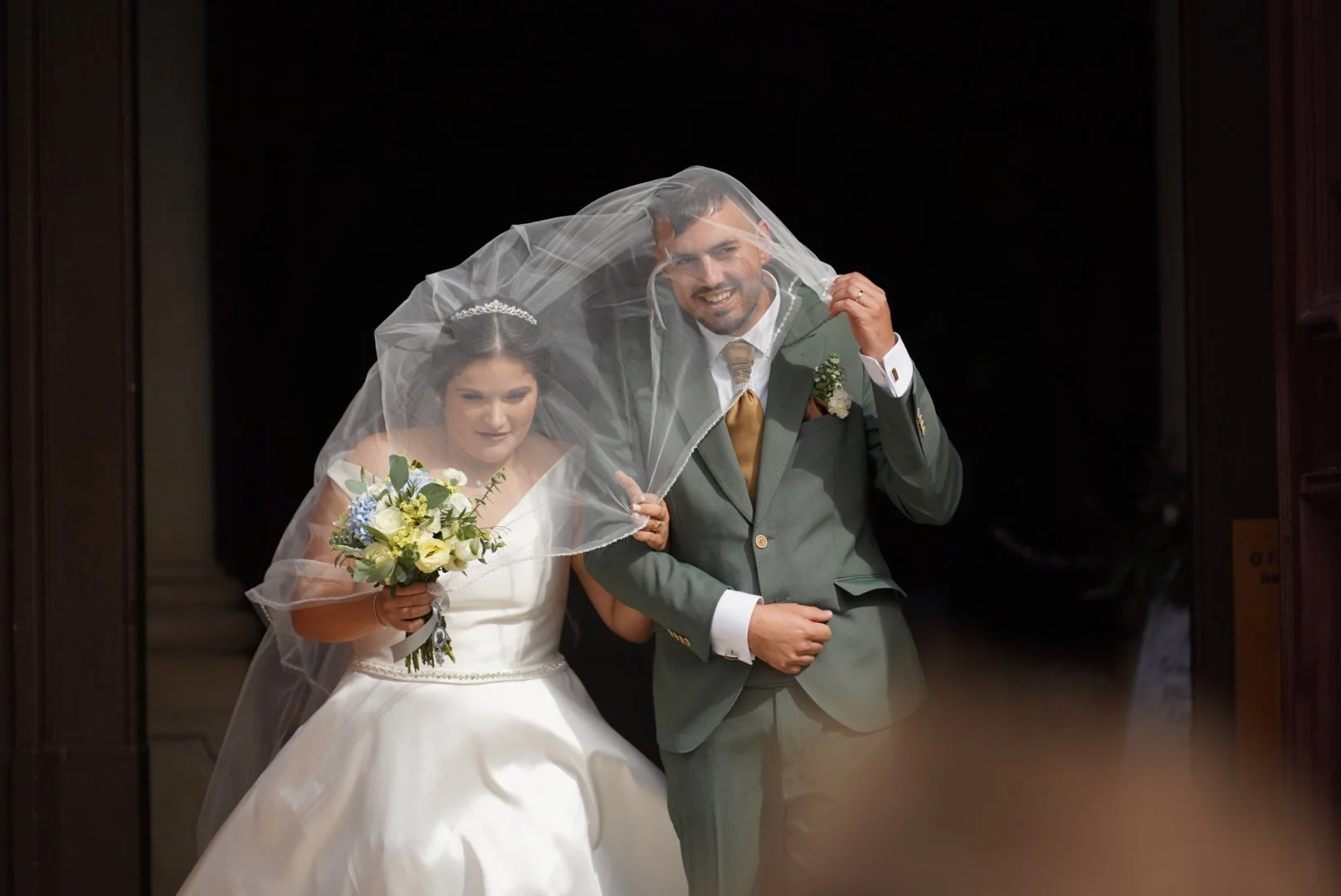 A bride and groom smiling together, with the groom holding a veil over the bride's head. The bride is holding a bouquet of white and purple flowers. They are dressed in wedding attire.
