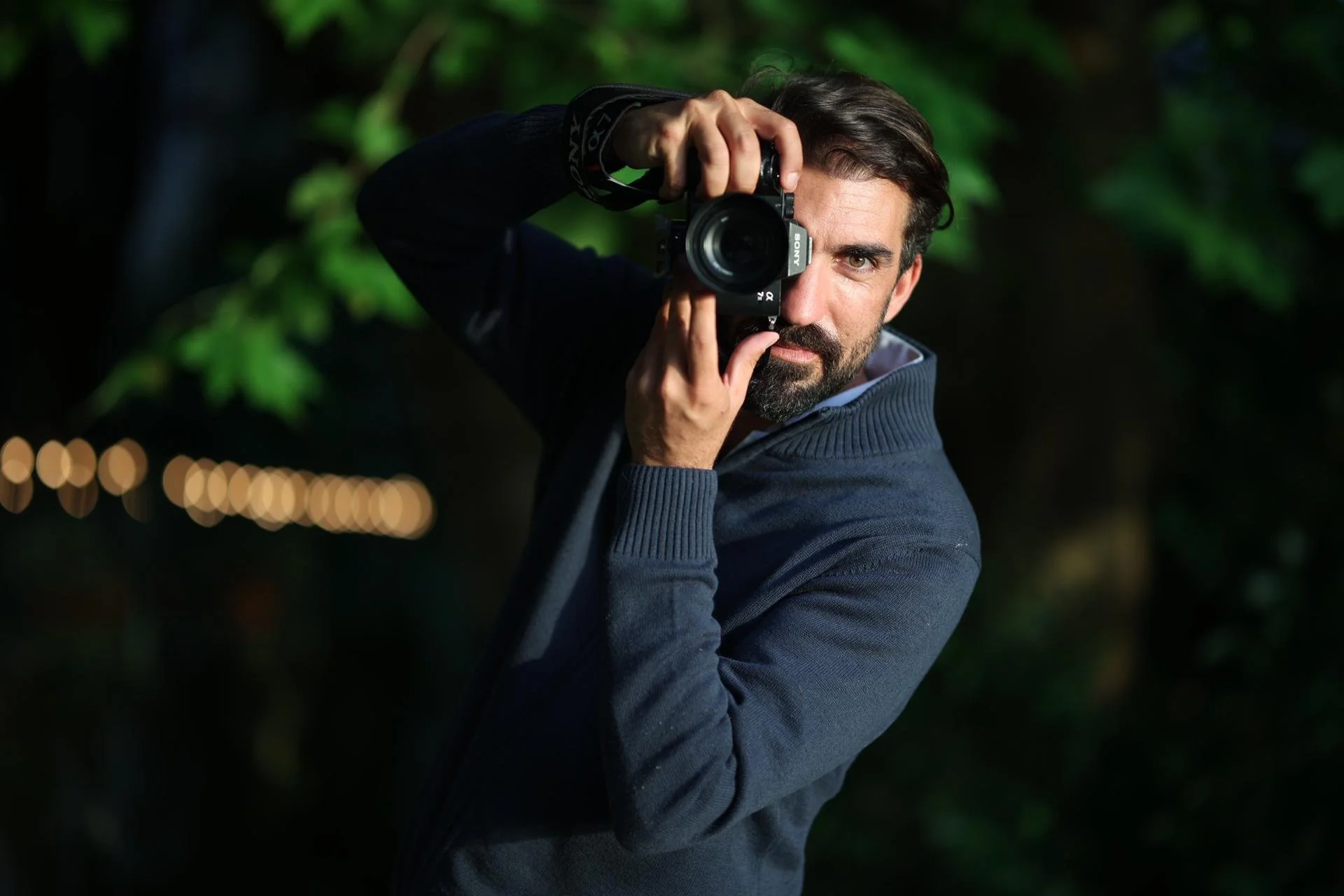 A man with dark hair and beard holding a professional camera up to his face, taking a photo outdoors at night with blurred lights in the background.