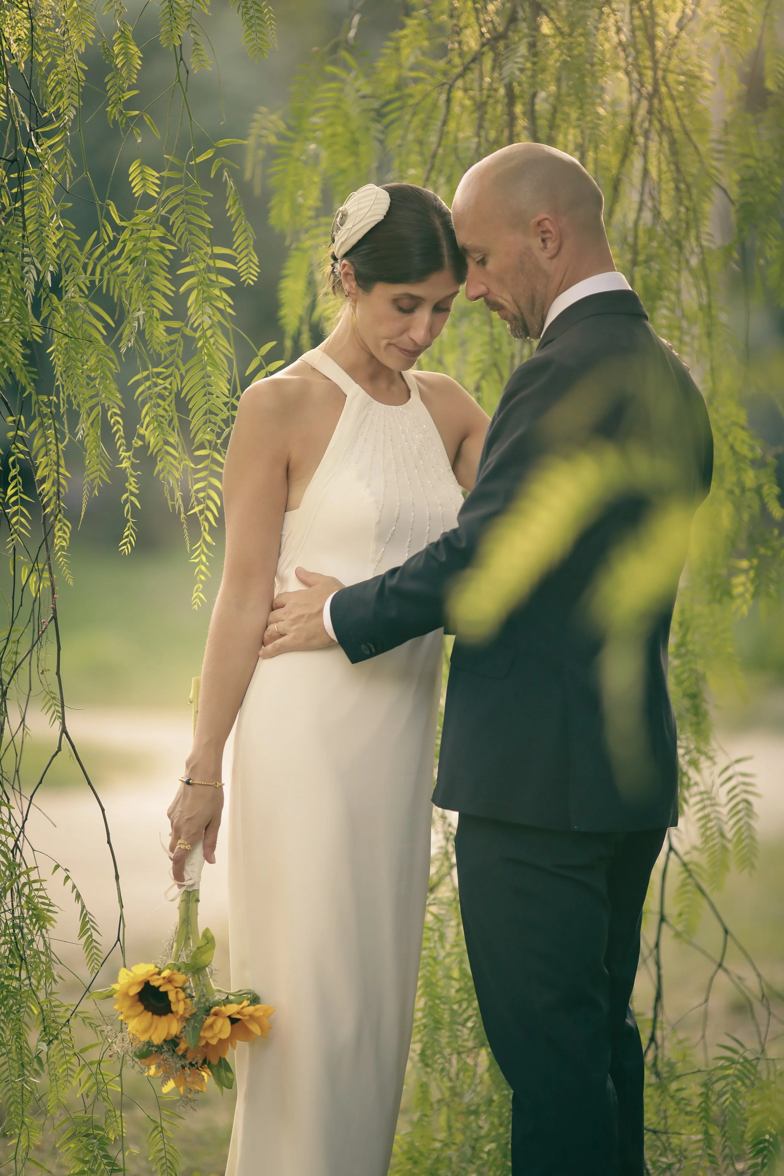 A couple dressed in wedding attire standing close together outdoors, surrounded by green hanging foliage, with a bouquet of sunflowers hanging at the woman's side.