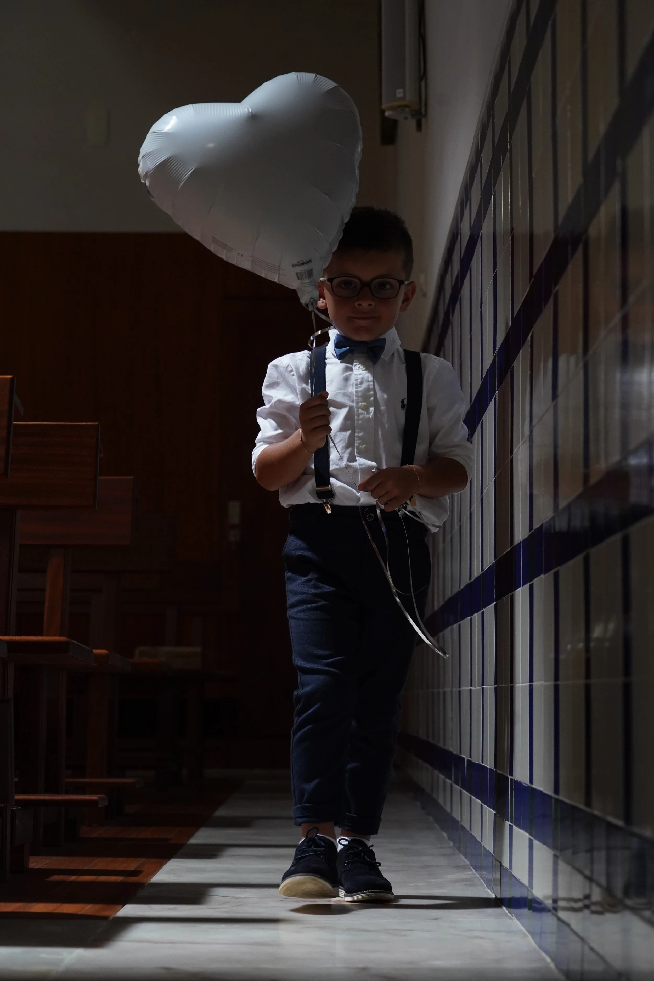 A young boy with glasses, dressed in a white shirt and black suspenders, holding a white heart-shaped balloon, walking along a tiled wall corridor with wooden seating on the side.