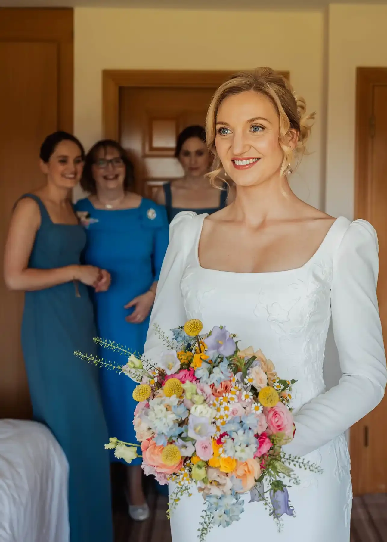 A woman in a white dress holding a colorful flower bouquet, smiling, with three women in blue dresses in the background.