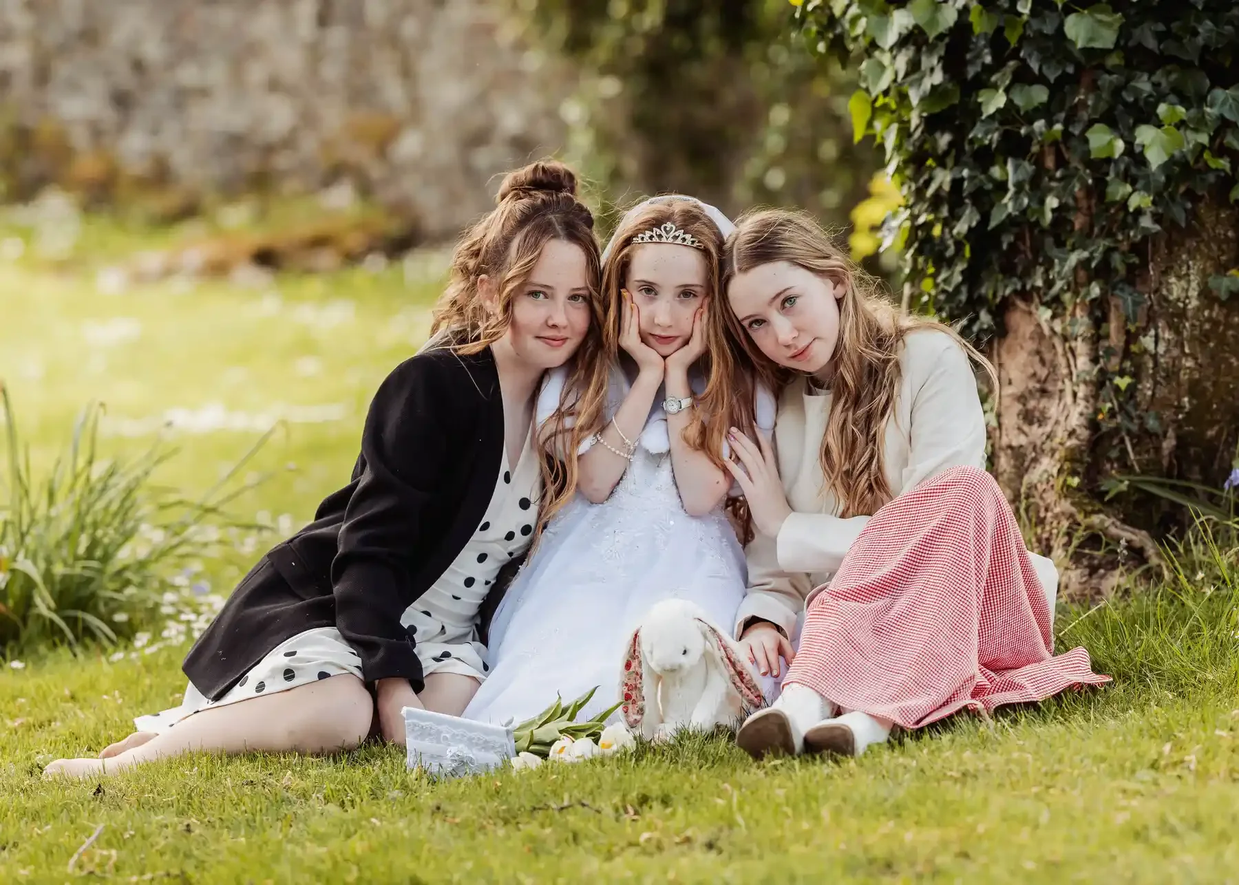 Three young girls sitting on the grass outdoors, with a pond and trees in the background. The girl in the middle is dressed in a white dress with a tiara, and the two girls on either side are casually dressed, leaning in close to her. There are white flowers and a stuffed animal on the ground in front of them.