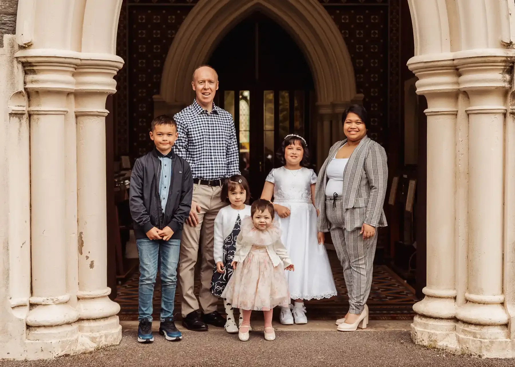 A group of seven people, including two men, one woman, and four children, standing on the steps of a historic building with stone columns and an arched doorway.