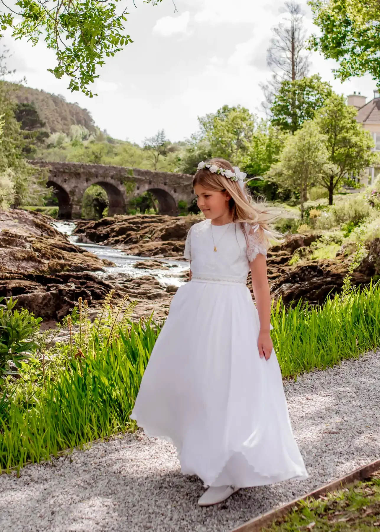A young girl in a white dress and floral headband standing near a stream with an old stone bridge in the background, surrounded by green trees.