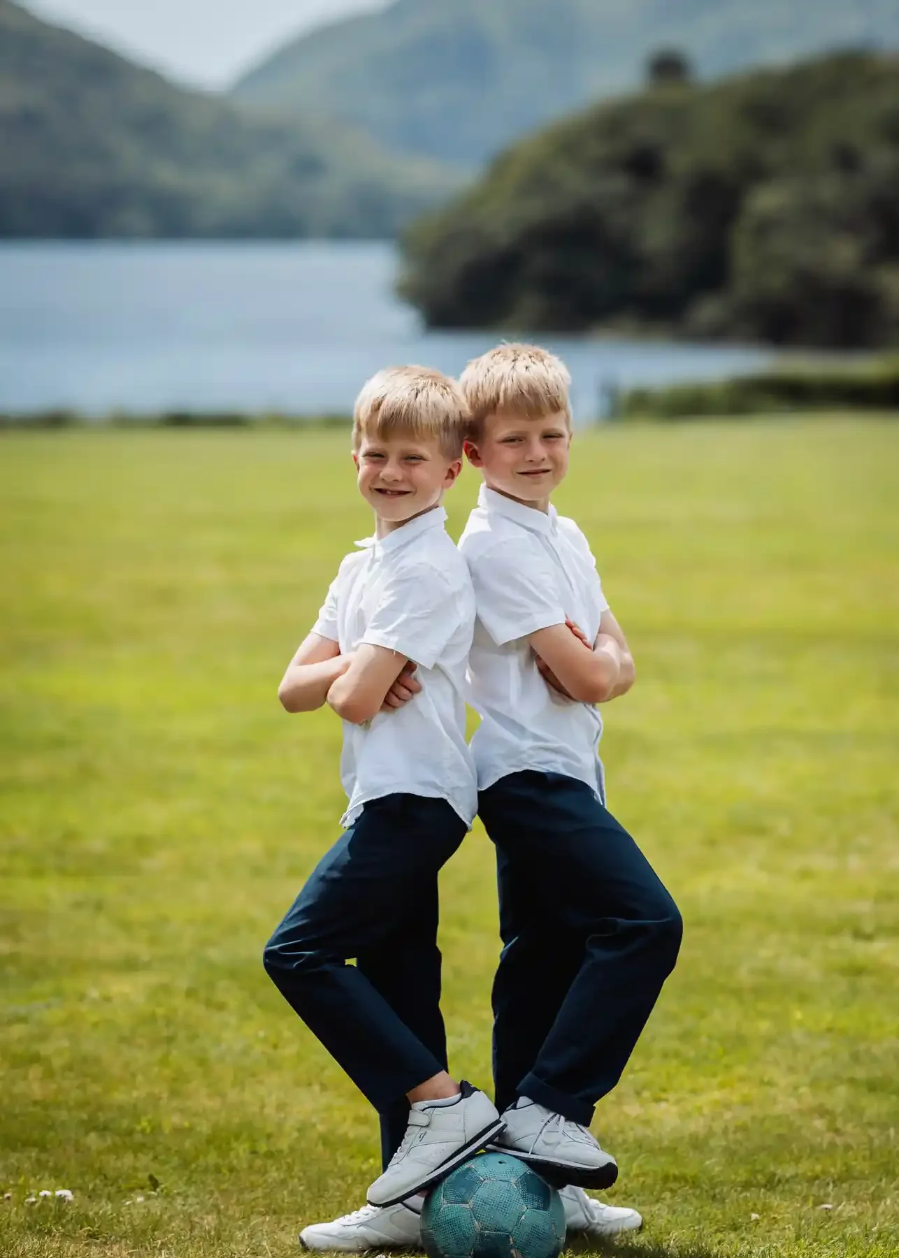 Two young boys with blond hair and white shirts standing back-to-back in a grassy field, with one foot on a soccer ball, smiling, with a lake and mountains in the background.