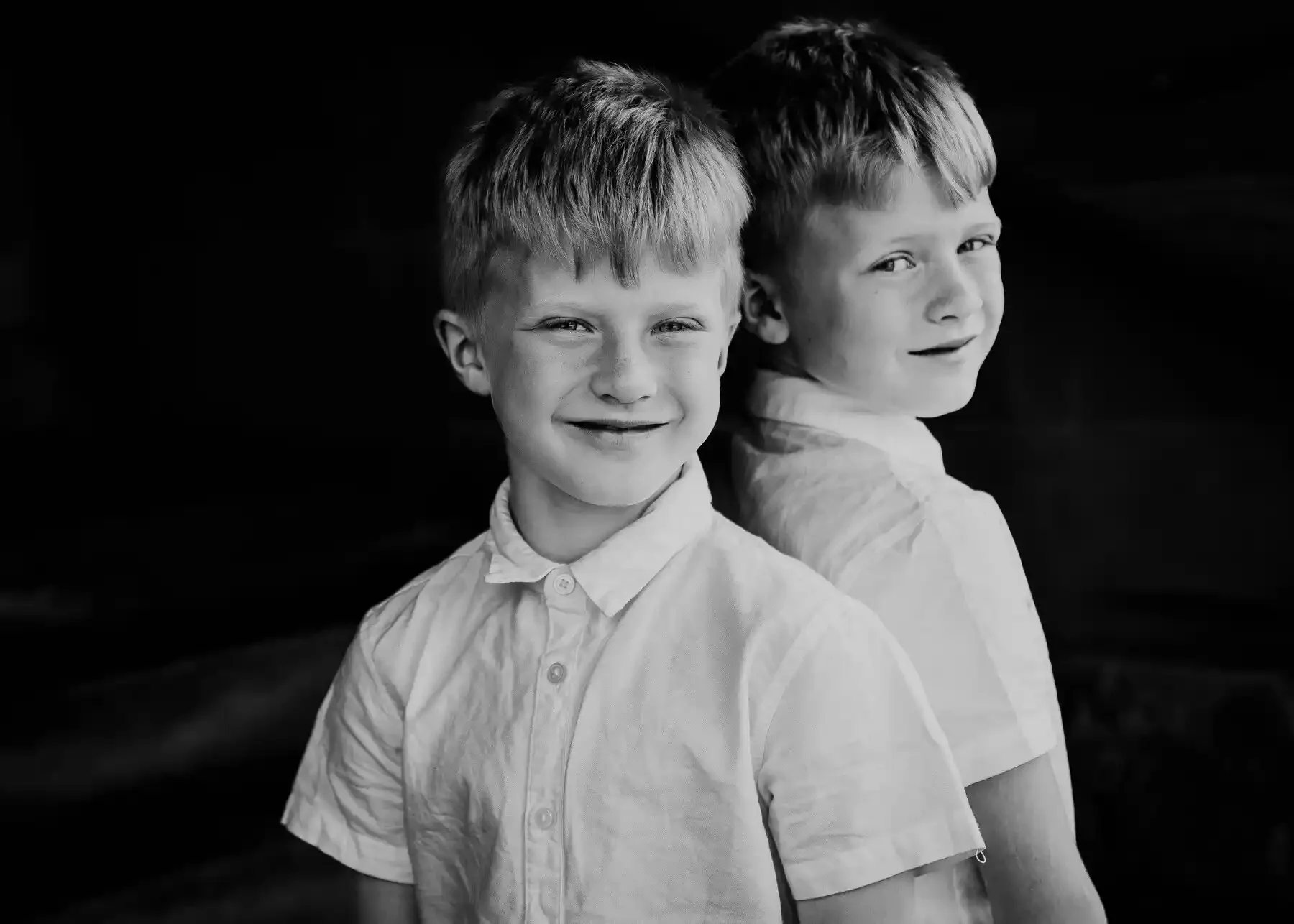 Two young boys in light-colored shirts stand back to back, looking at the camera. The boy in front is smiling, while the boy behind has a more neutral expression. The background is dark.