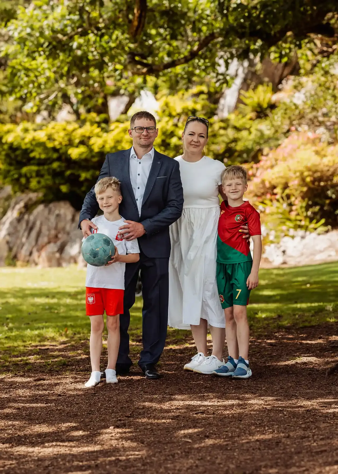 Family of four standing outdoors on grass with trees in the background, two young boys and two adults, one man in a suit and one woman in a white dress, the boys are in sports outfits, one holding a soccer ball.