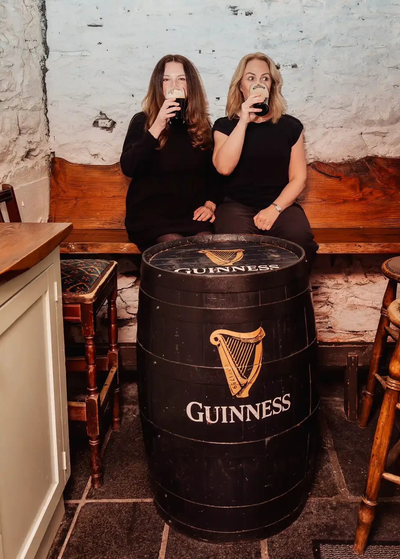 Two women sitting on a wooden bench in a rustic bar holding pints of dark stout beer, with a Guinness-branded barrel table between them, and white brick walls in the background.