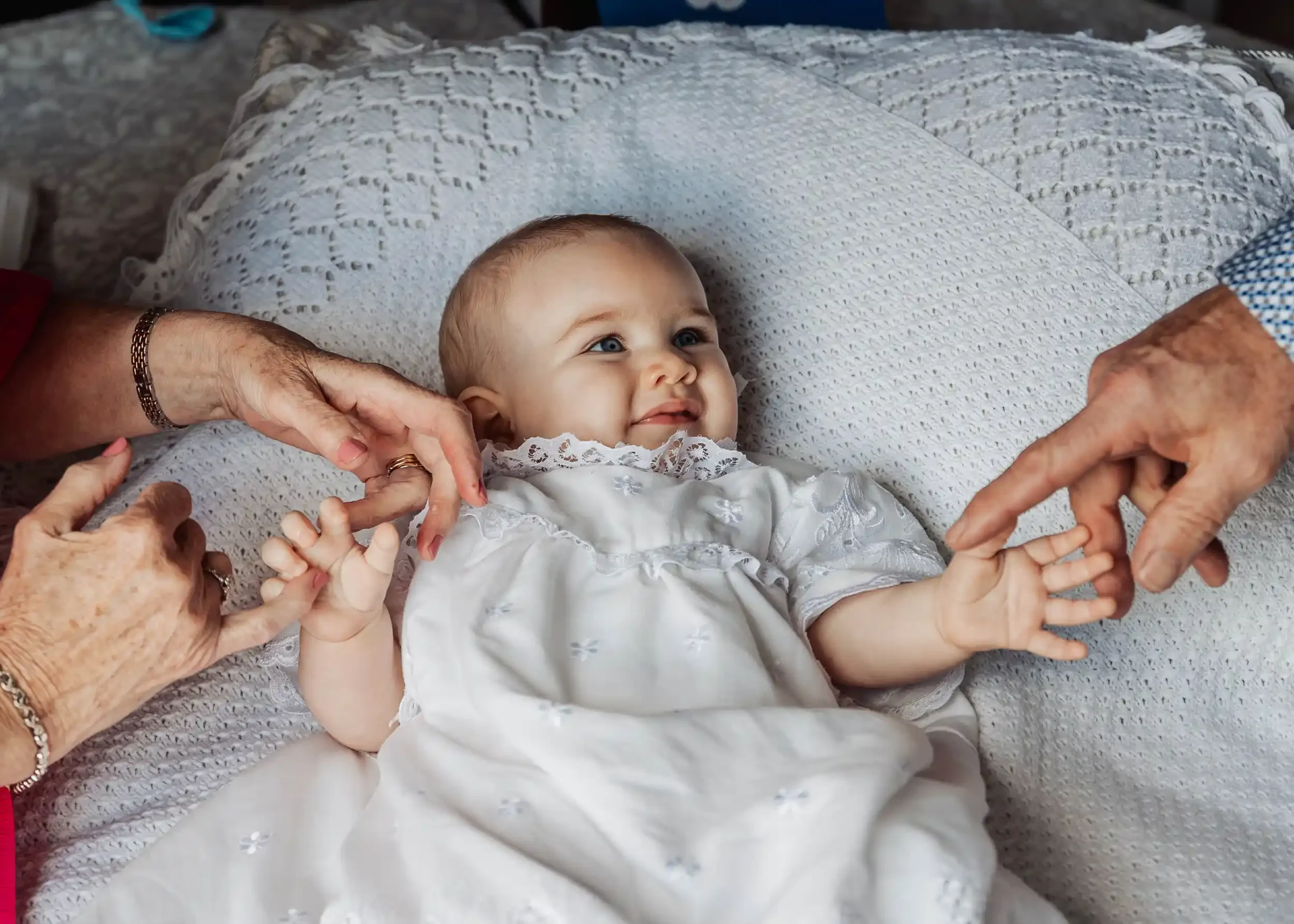 Baby lying on a bed, smiling, with elderly hands holding and gently touching the baby's hands.
