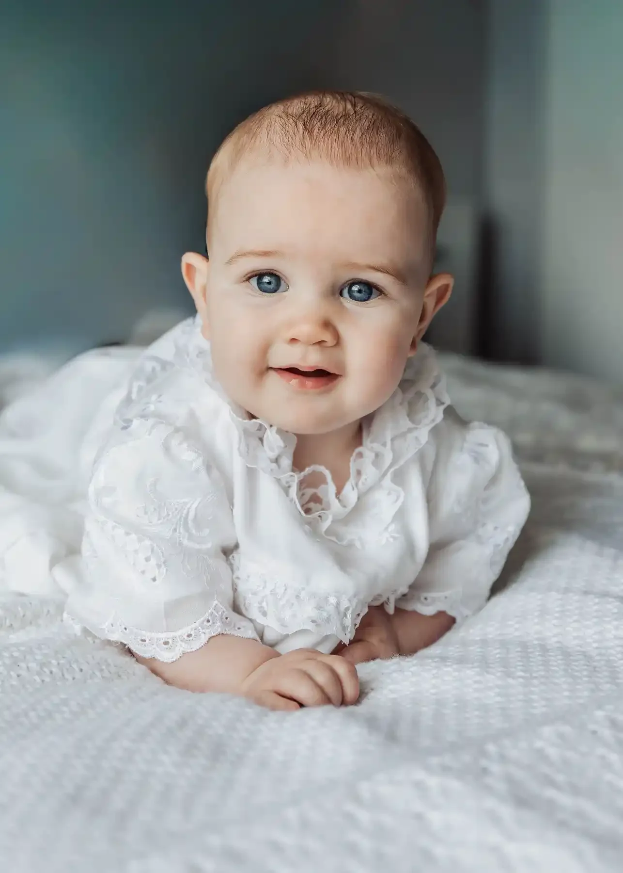 A young baby with blue eyes and light brown hair, wearing an elegant white lace outfit, lying on a soft white blanket, looking at the camera with a gentle smile.