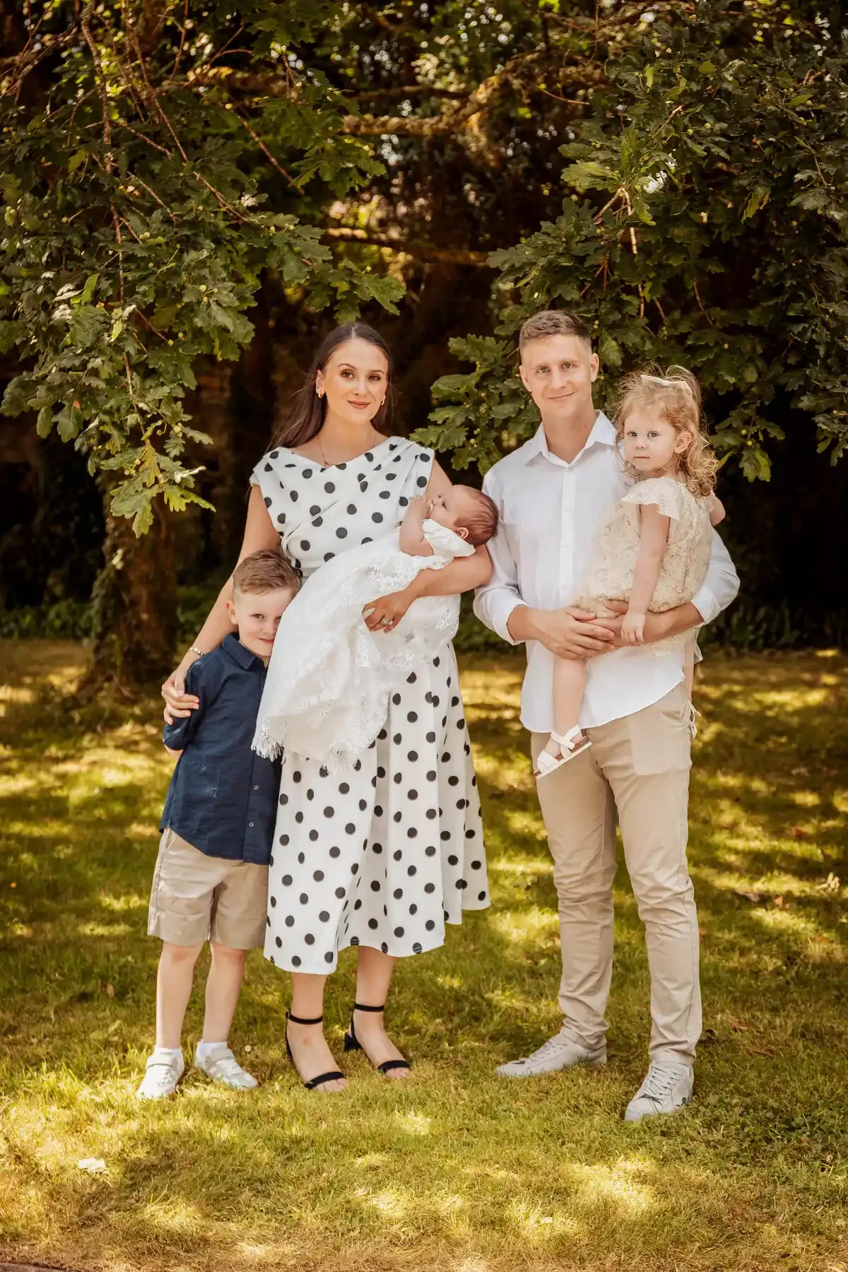 A family of five posing outdoors in front of a large leafy tree during daytime. The mother holds a newborn baby dressed in white, and stands next to a young boy in a navy shirt and khaki shorts. The father holds a young girl in a light dress, with al