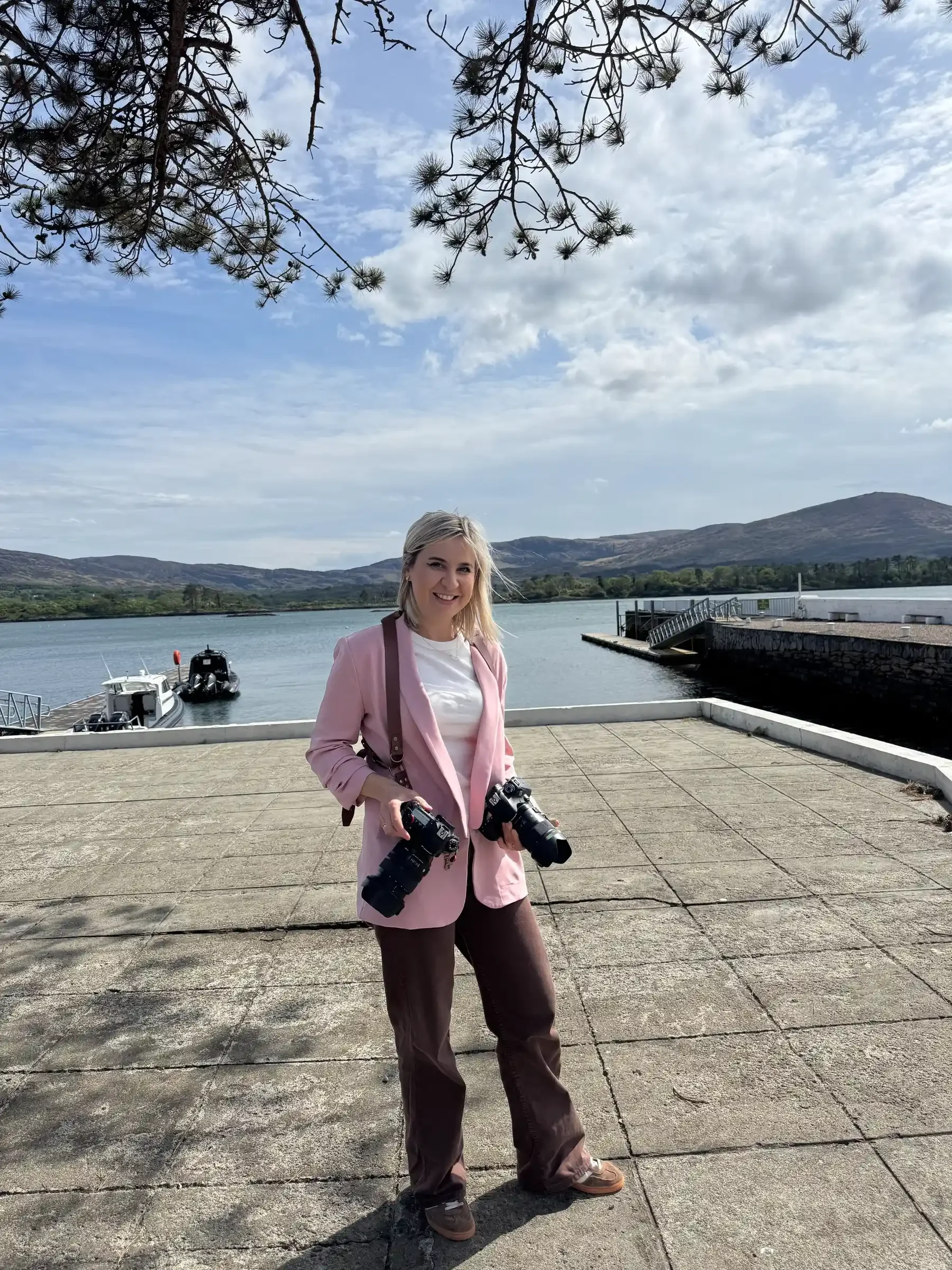 A woman standing on a waterfront promenade holding cameras with a lake, boats, trees, hills, and a partly cloudy sky in the background.