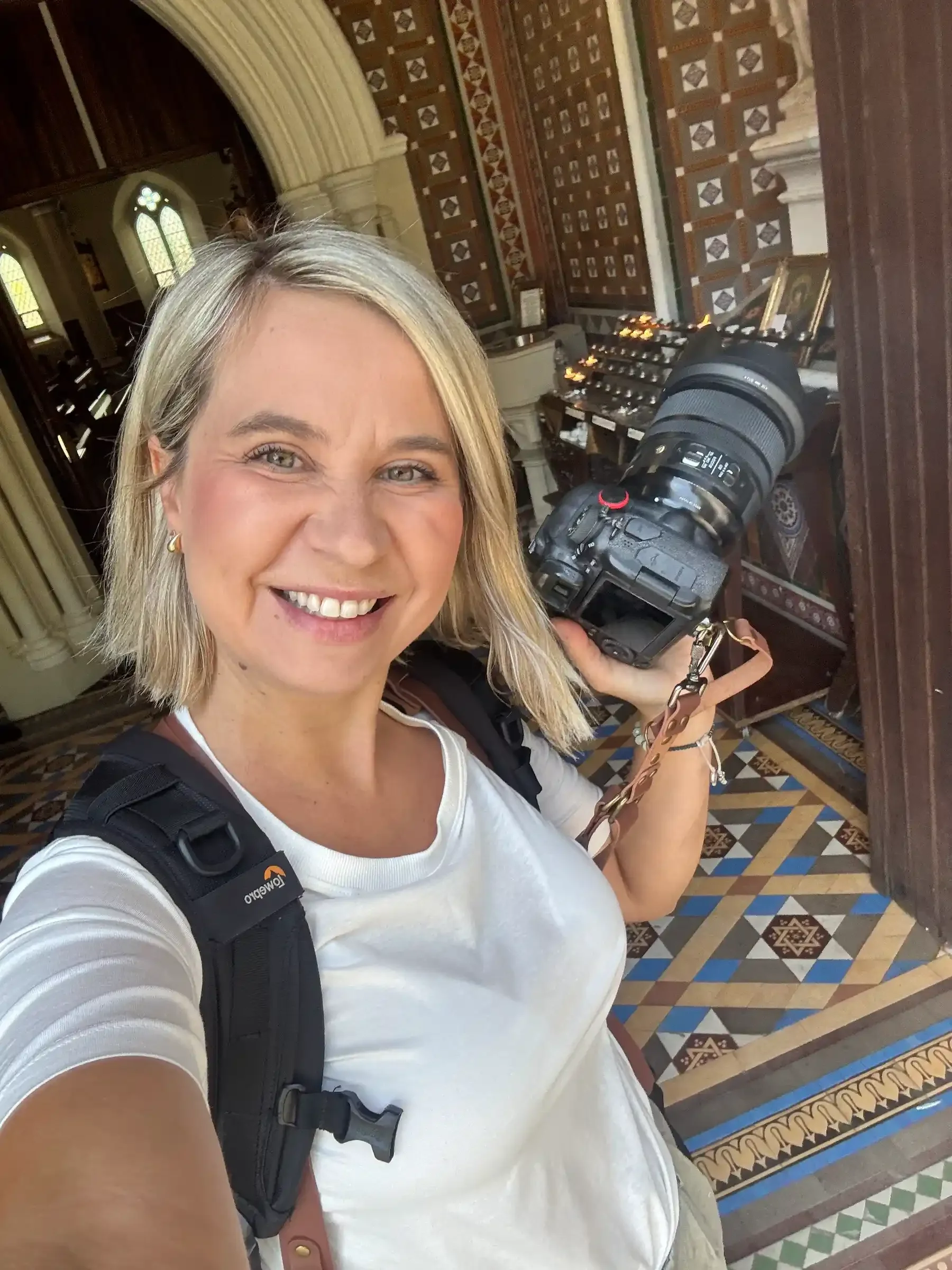 A woman with blonde hair smiling while holding a camera inside a building with ornate tiled floors and wood-paneled walls.