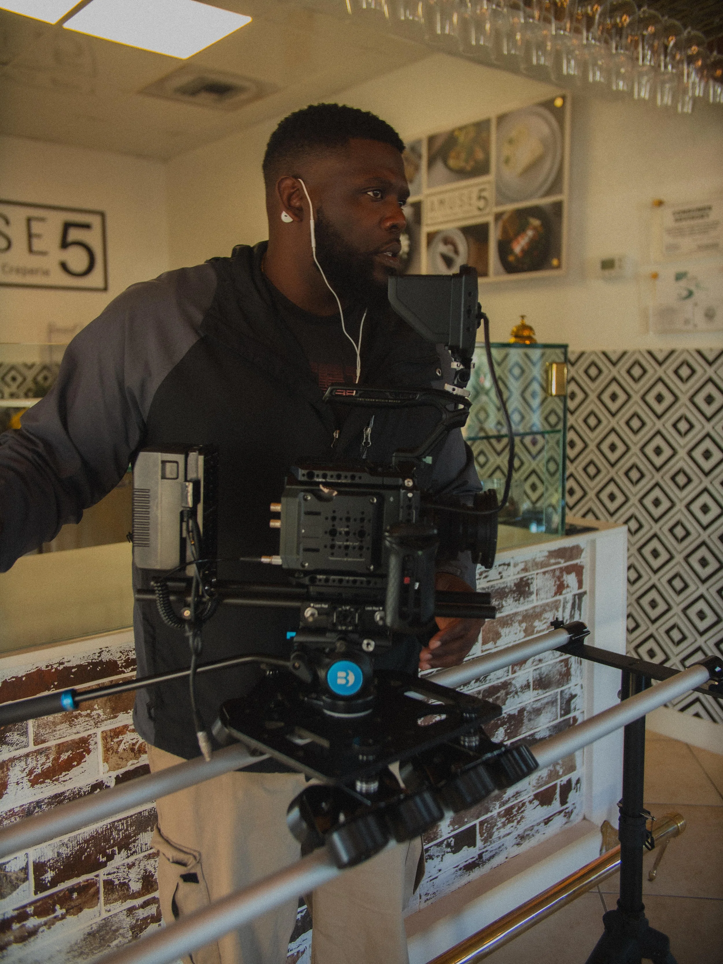 A man operating a professional camera rig on a tripod inside a restaurant or cafe, with decor including a sign that says 'Choose 5' and patterned tiles on the wall.