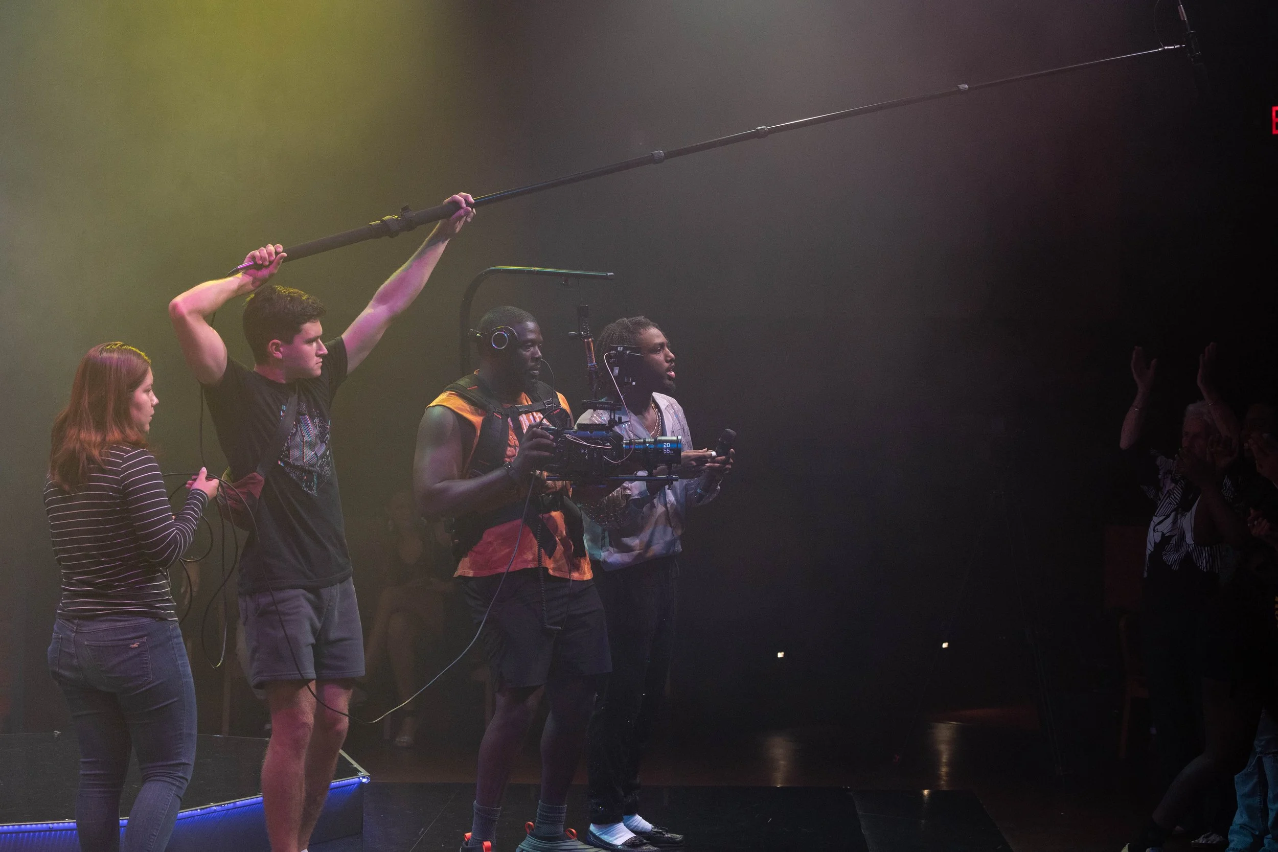 A film crew is filming a scene on a stage with dark lighting and fog, including a woman with red hair and a striped shirt, a man holding a boom microphone, and three other crew members with equipment.