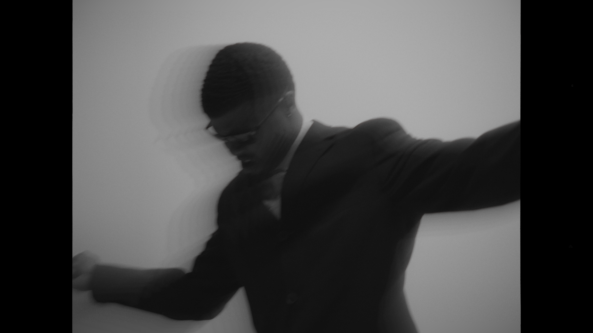 Black and white photo of a man in a suit leaning against a wall, wearing glasses and a hat.