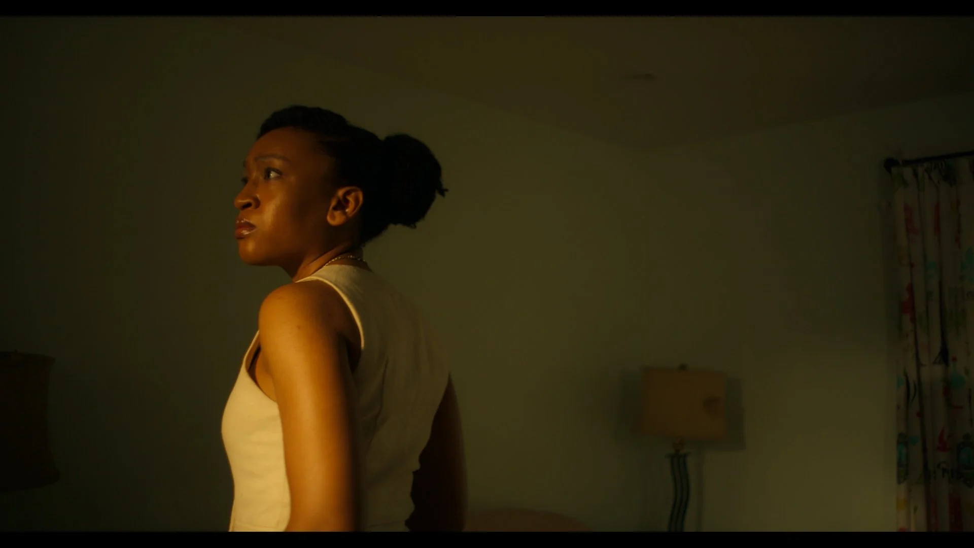 A woman with dark hair styled in an updo, wearing a beige top, stands in a dimly lit room, looking to her left with a serious expression. There is a beige lamp and a window with colorful curtains in the background.