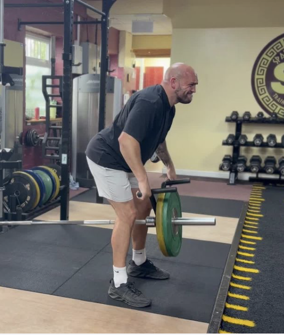 Man with a bald head and tattooed arm lifting a barbell loaded with colored weights in a gym, showing strain.