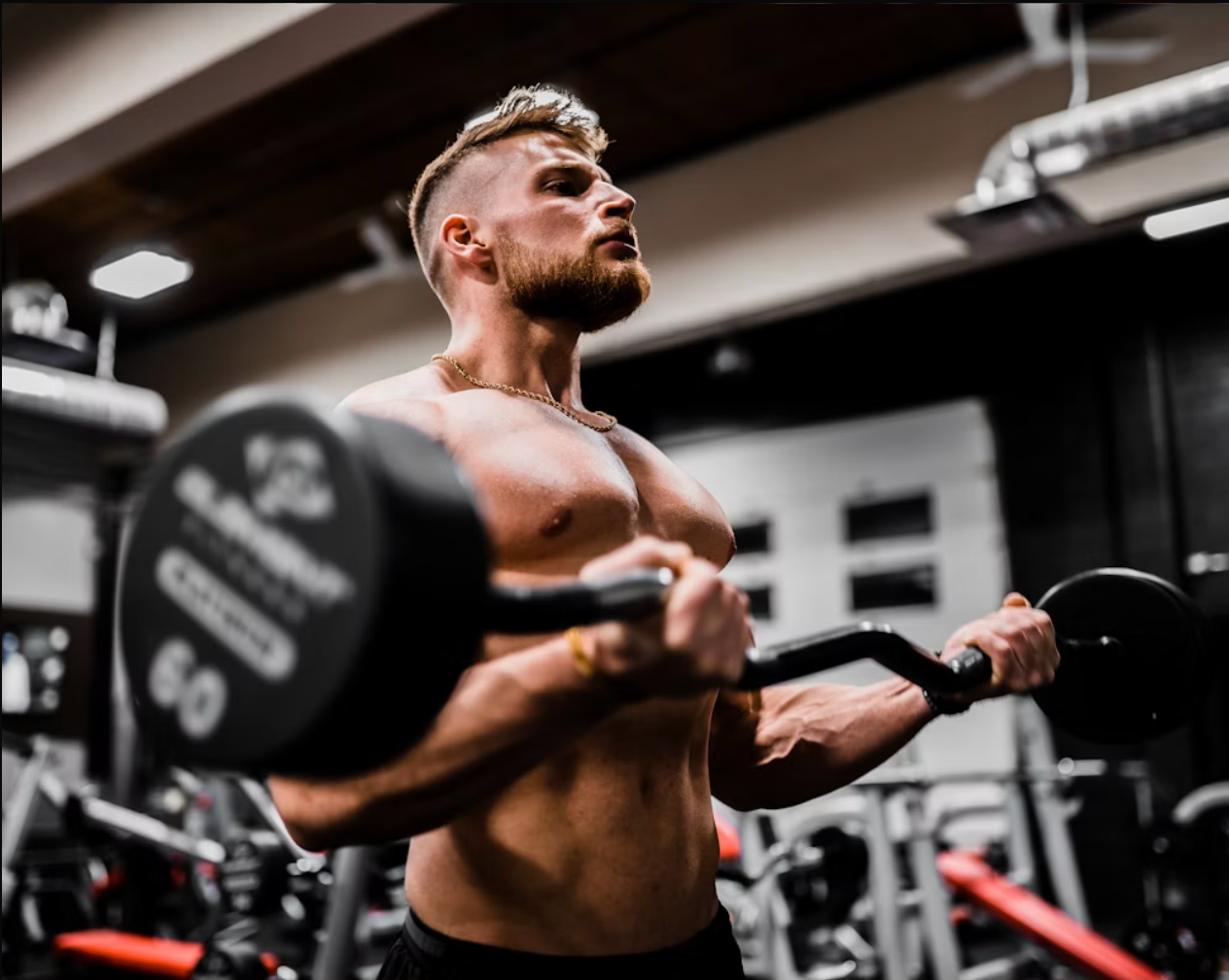 A muscular man with a beard and styled hair, shirtless, lifting a dumbbell in a gym.